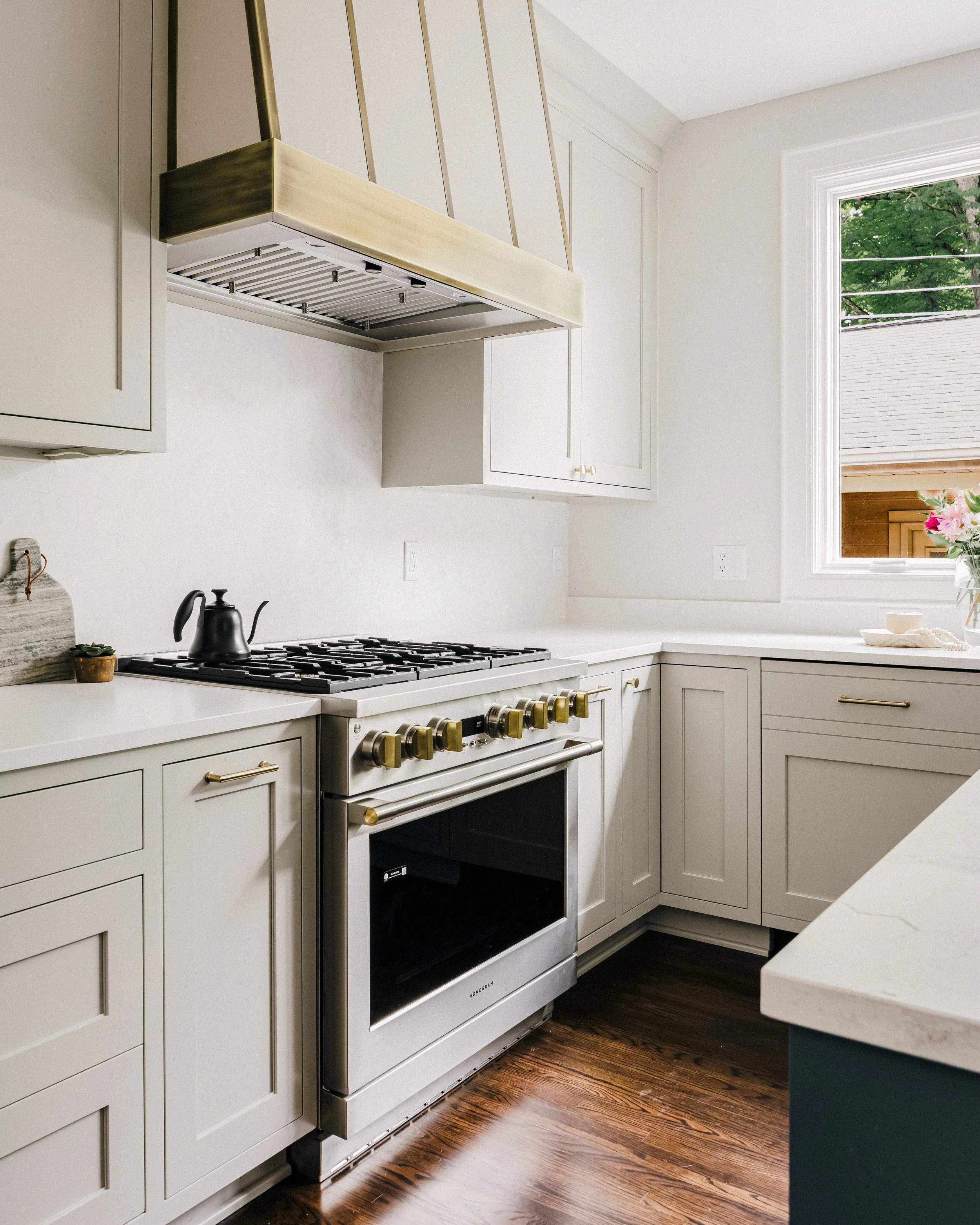 Bright kitchen with custom Maple cabinets, luxury Monogram range with gold accents, and brass strapping on a Maple hood. A teapot sits on the stove. Sunlight filters through a window.