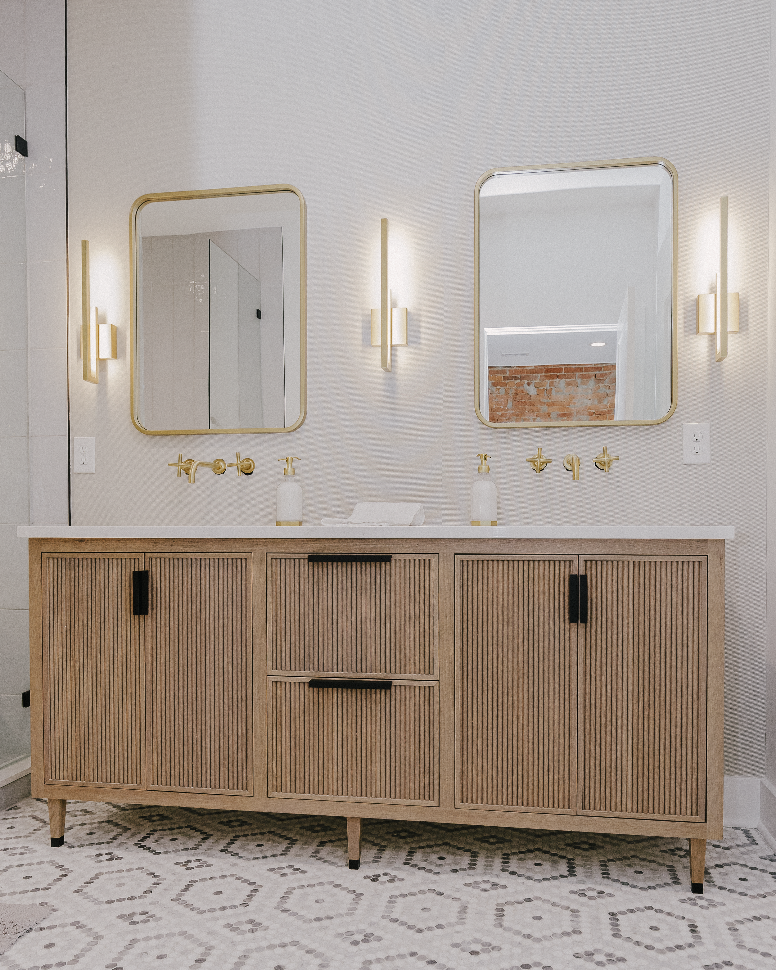 Modern bathroom with a reeded vanity cabinetry, dual gold wall-mounted faucets, and chic sconces flanking two gold-rimmed mirrors. Bright, clean aesthetic conveyed by patterned tile flooring and soft lighting.
