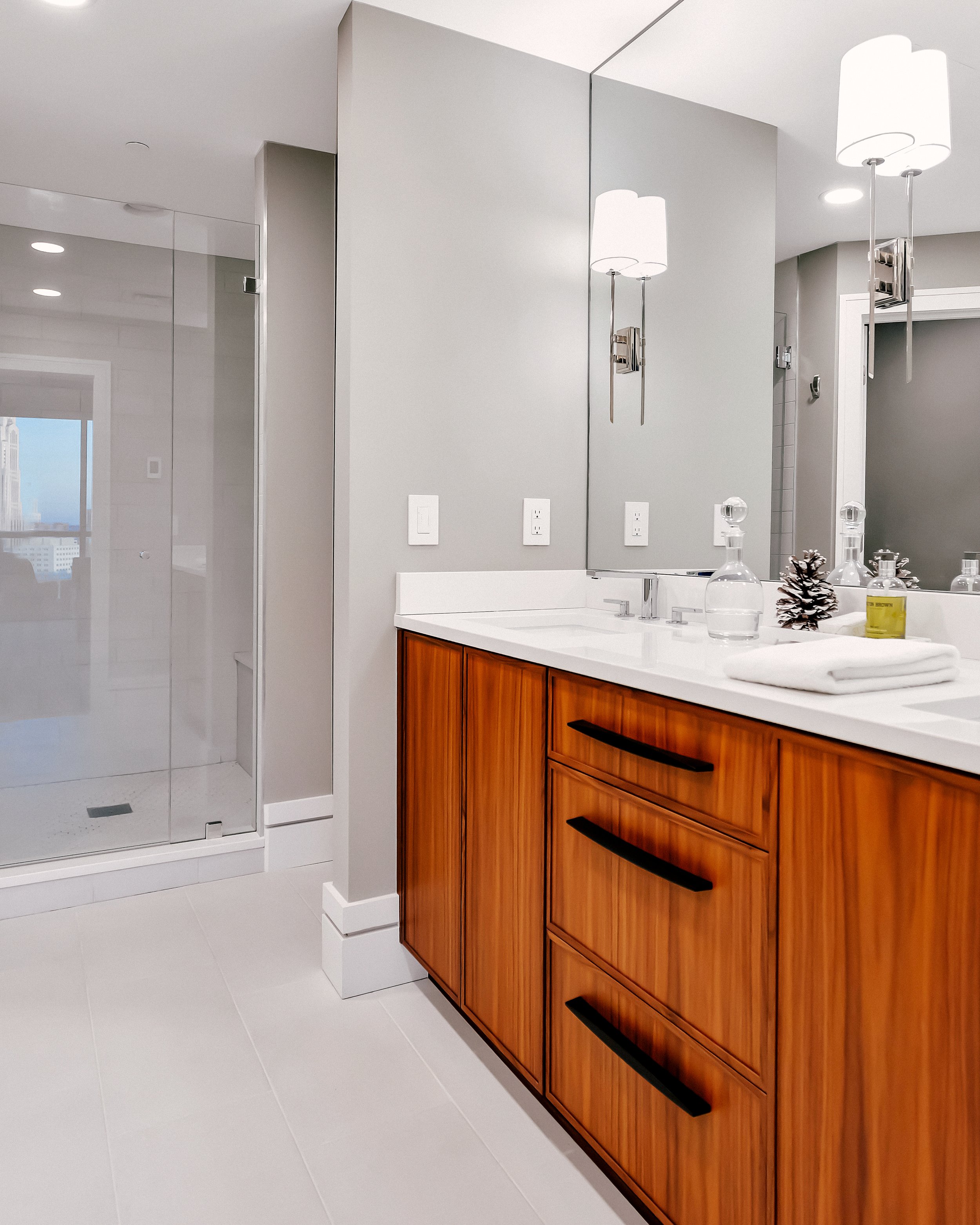 Modern bathroom with a custom wooden vanity accented with matte black handles, a large mirror, and soft lighting. A frameless shower peeks into the photo.