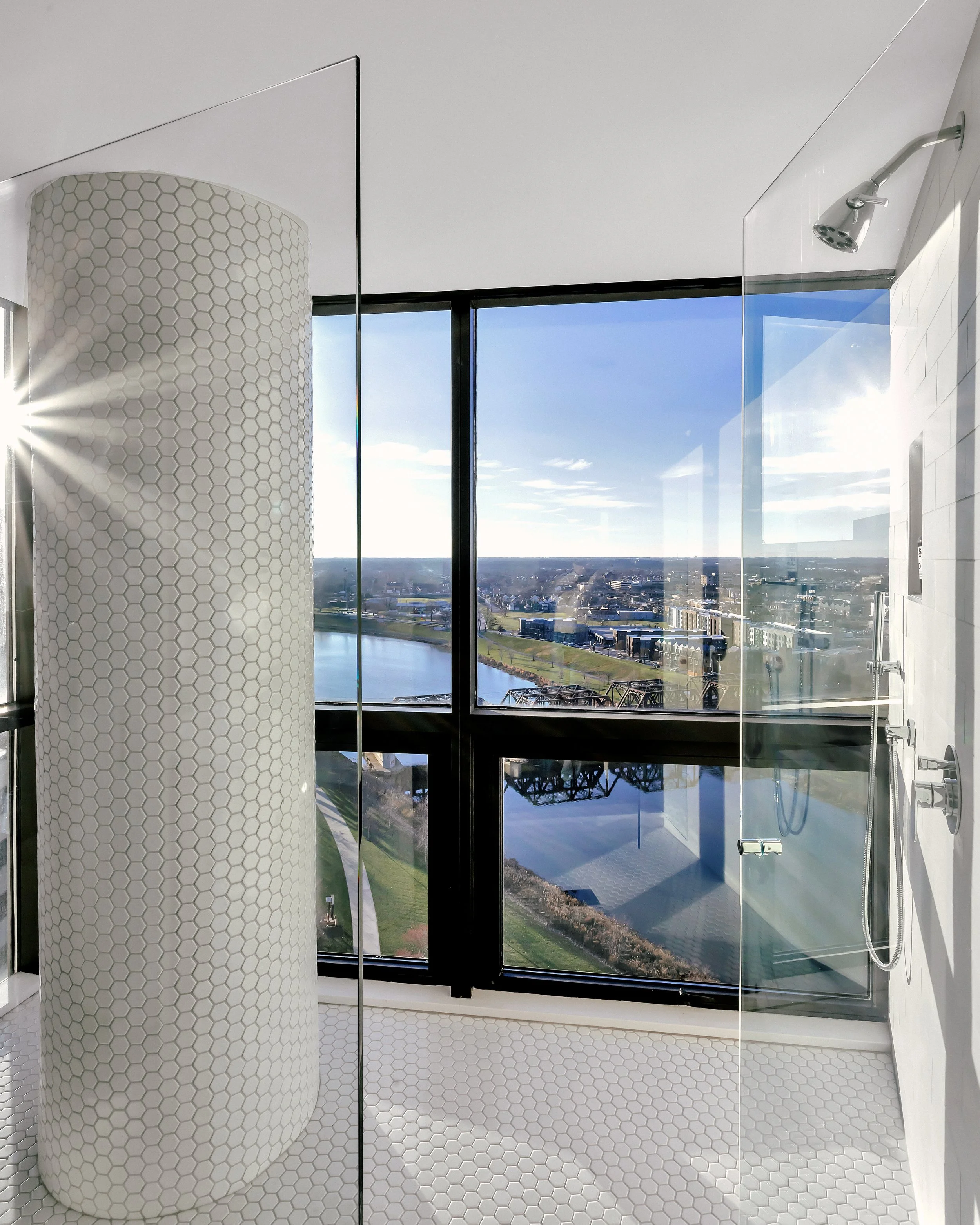Modern bathroom with glass-walled shower and hexagonal tiles, overlooking the Columbus Scioto River through large windows. Bright and serene ambiance.