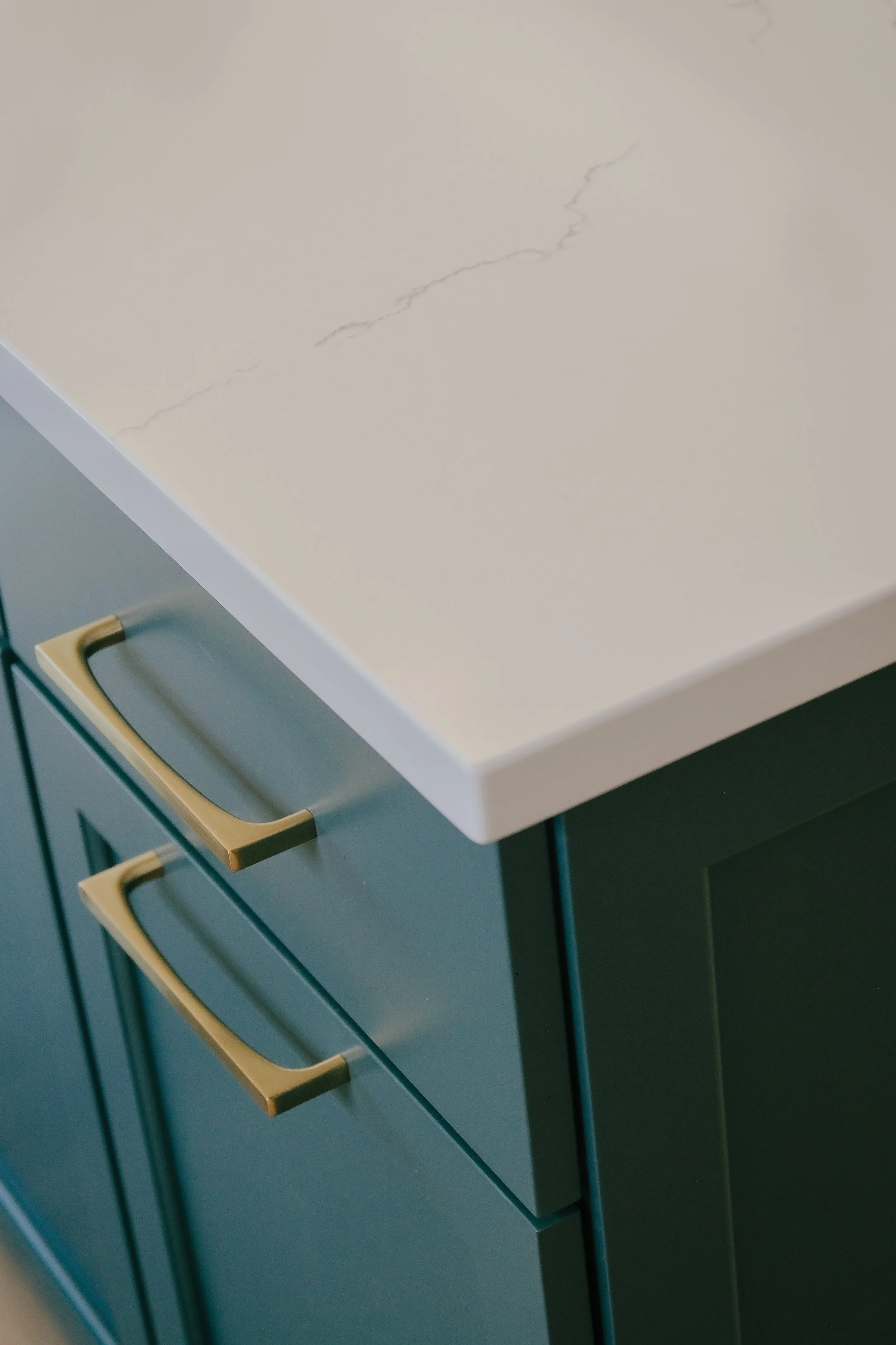 Close-up of a kitchen island with a smooth, white Cambria quartz surface featuring subtle veining. Dark green cabinets with sleek gold handles below.