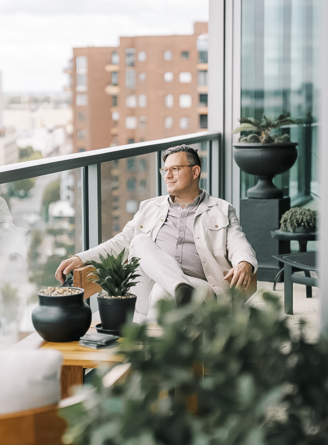 Tyler Swartzmiller, owner and lead interior designer of Haus Studio, in a light jacket sits relaxed on a modern balcony with city views, surrounded by potted plants. The scene conveys tranquility and urban charm.
