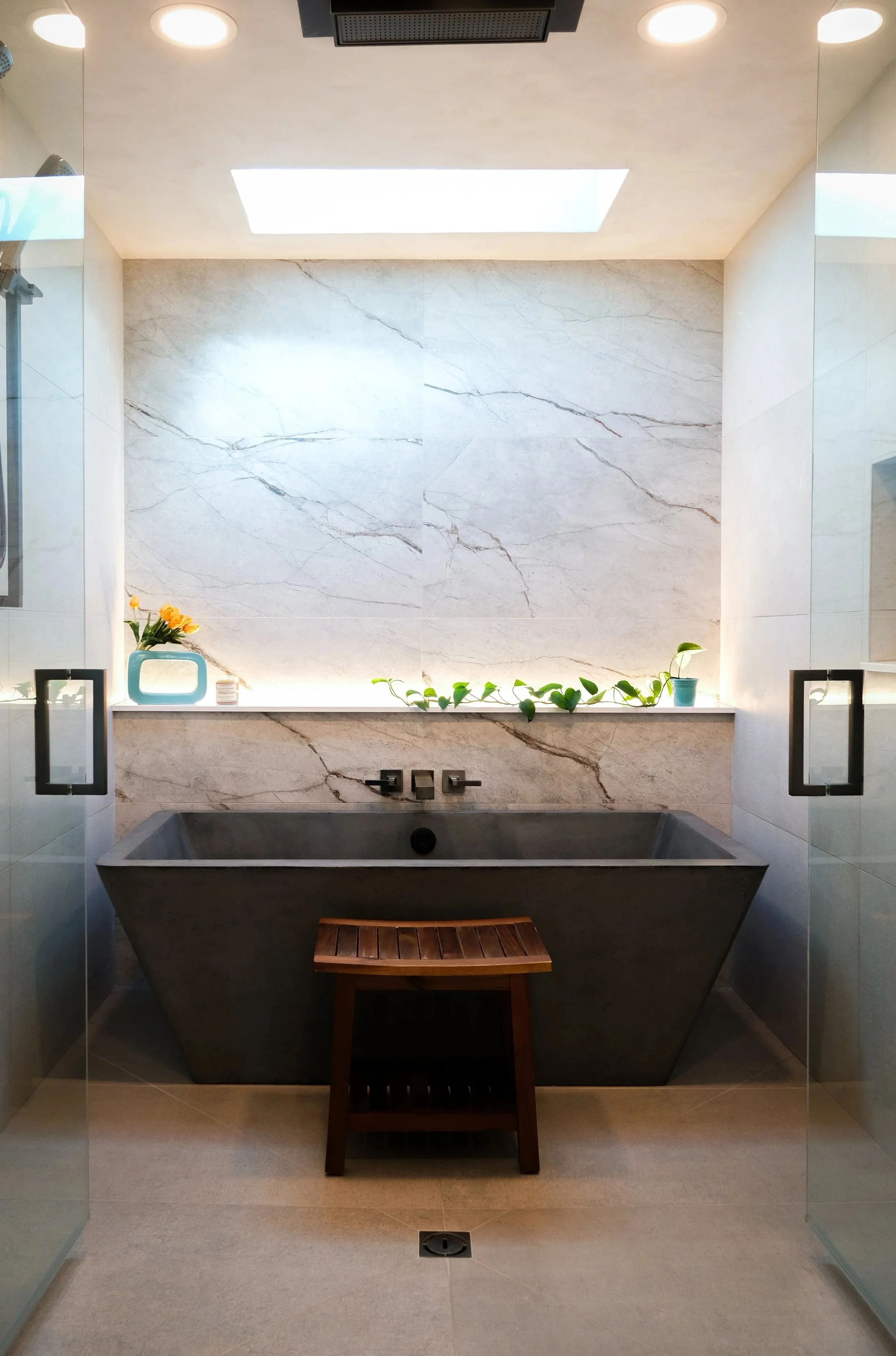 A stunning full-height stone statement wall overlooks a concrete in-shower tub next to a wooden shower bench in this moody bathroom. A skylight illuminated the shower space.