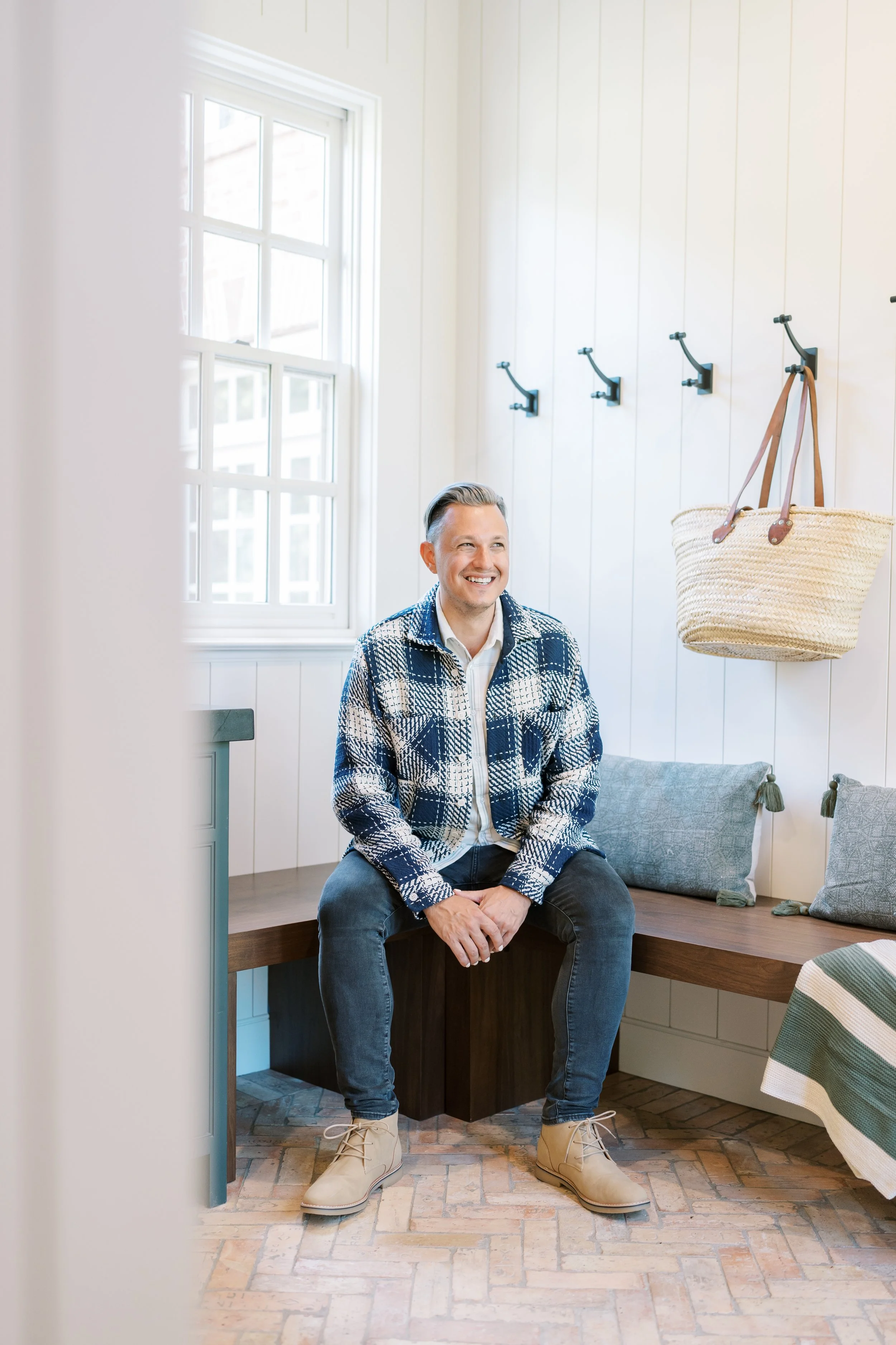 Tyler Swartzmiller, owner and lead designer at Haus Studio, sits on a wooden bench in a neutral, airy mudroom.