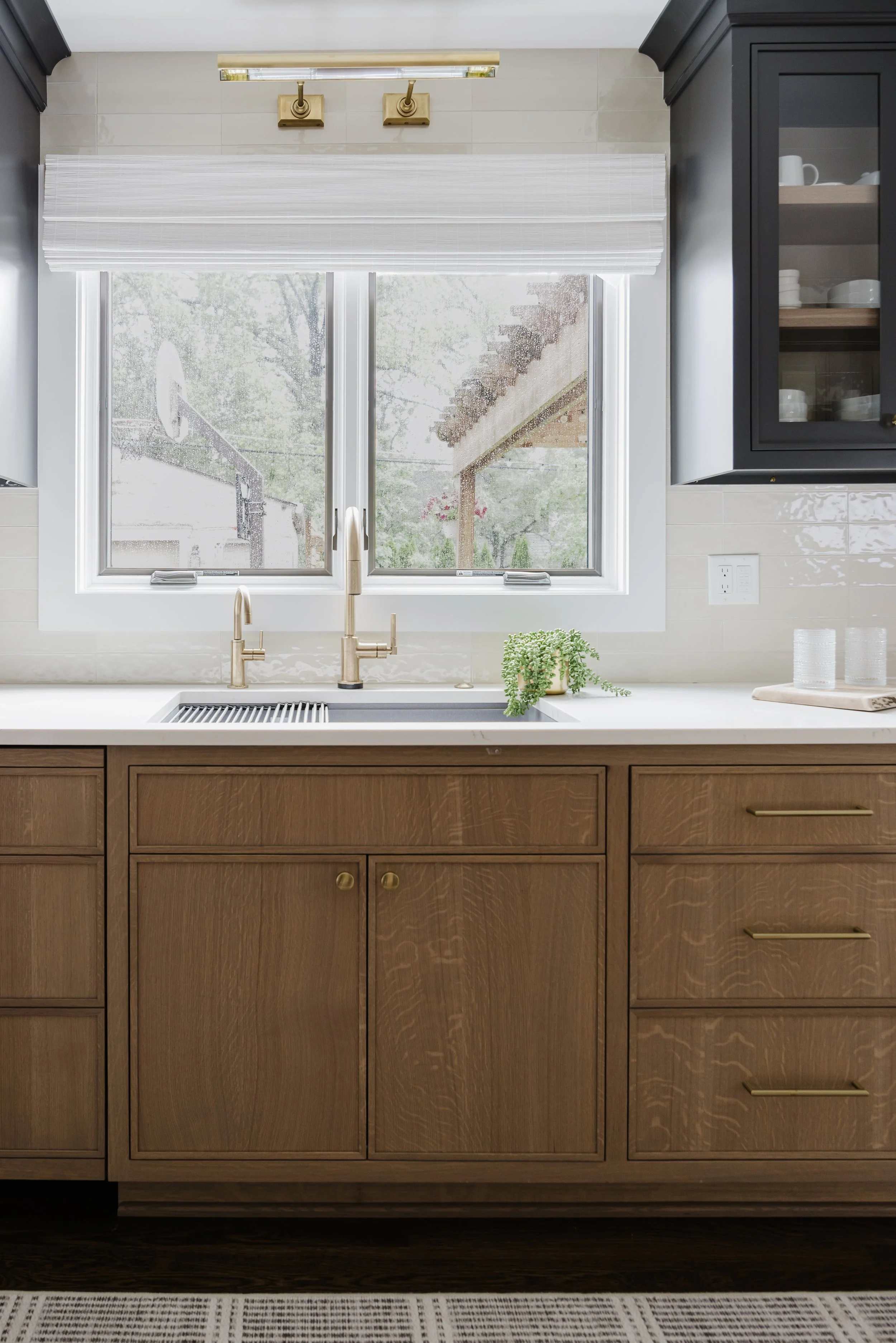 Modern kitchen with White Oak cabinets, a Cambria quartz countertop, and Brizo faucet centered under a window. Natural light and greenery add freshness.
