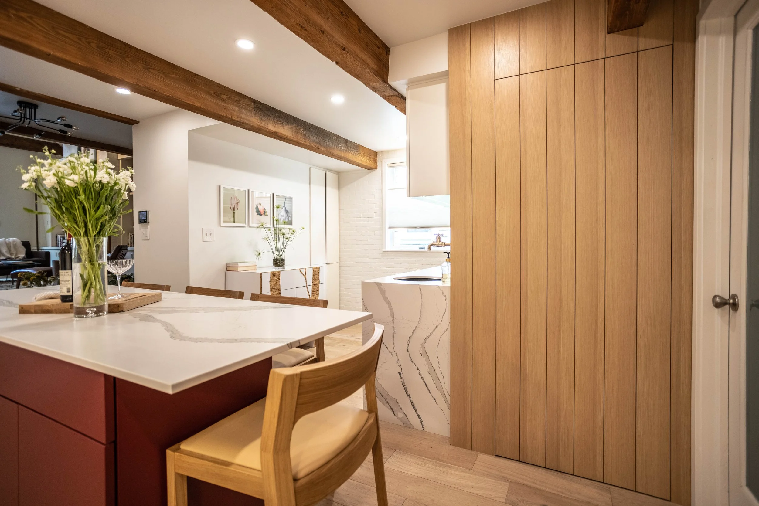 Scandinavian kitchen with exposed wooden beams and gorgeous red island with white Cambria quartz countertop, light wooden chairs, and bright, airy atmosphere. A paneled White Oak wall hides a door to kitchen storage.