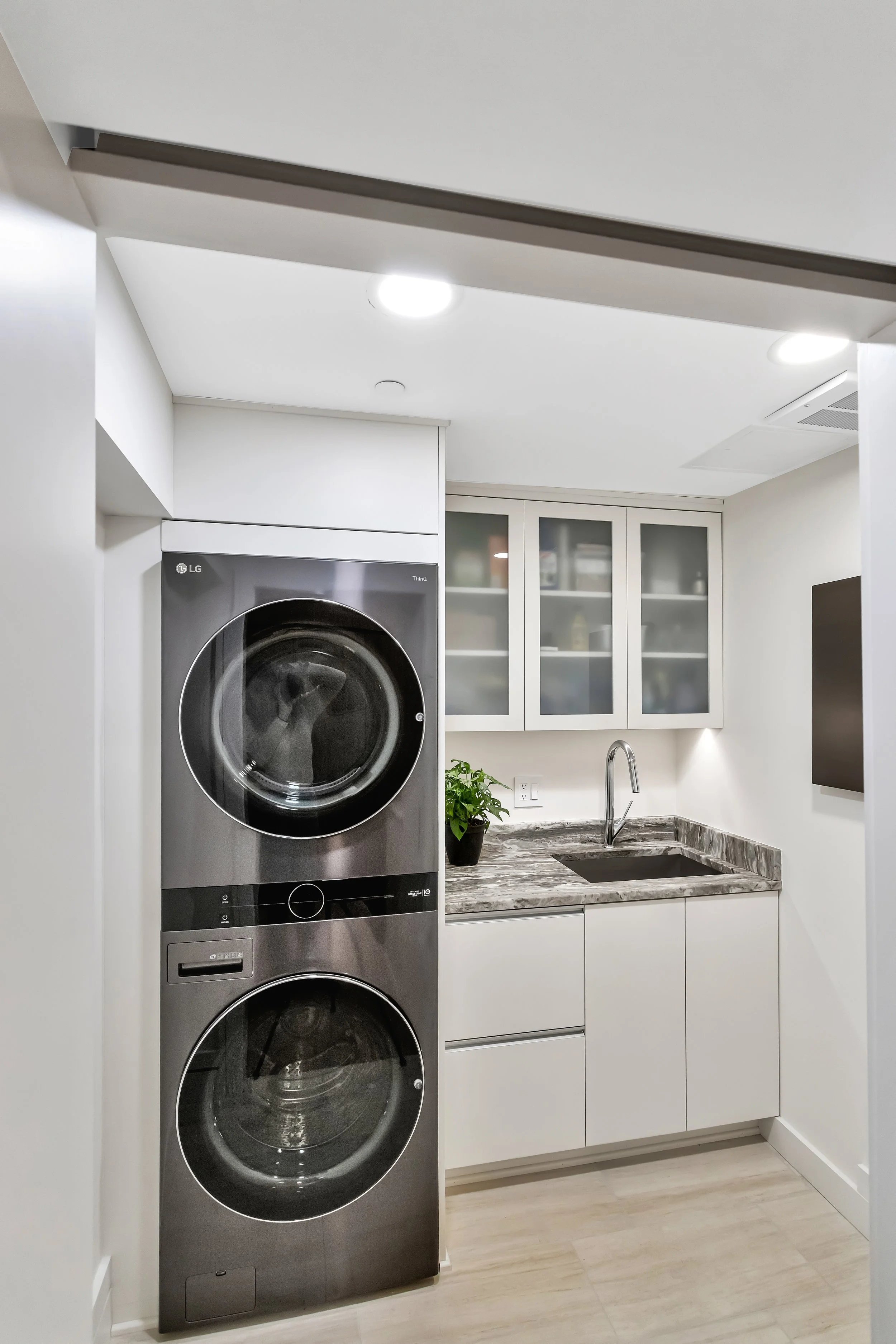 Laundry room with a sleek, stacked washer and dryer on the left, a quartz countertop with a sink, white frosted cabinets above, and bright lighting.