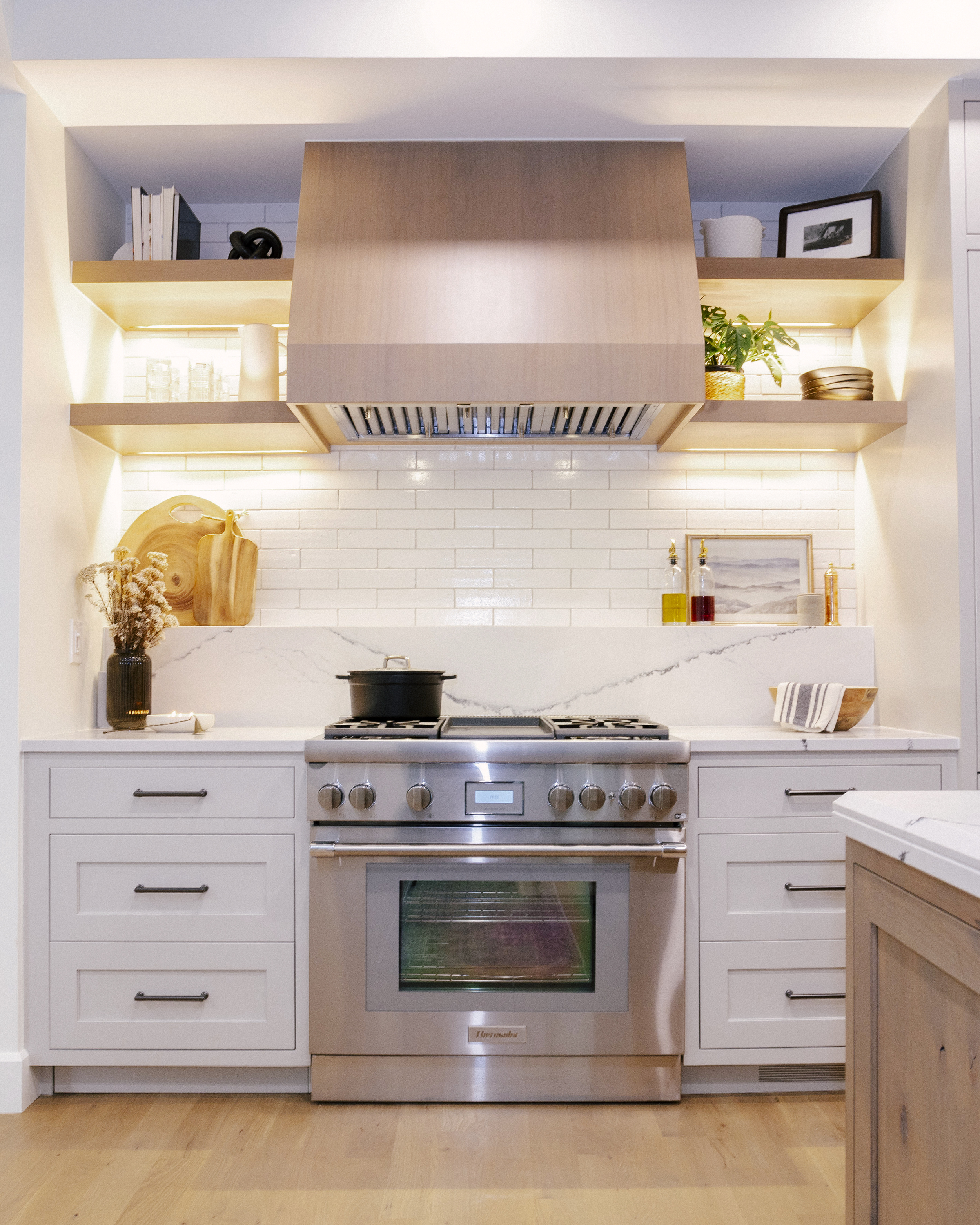 Modern kitchen with a Thermador range, quartz countertop, and mixed quartz and tile backsplash. Warm lighting highlights a bold White Oak range hood and wooden shelves.