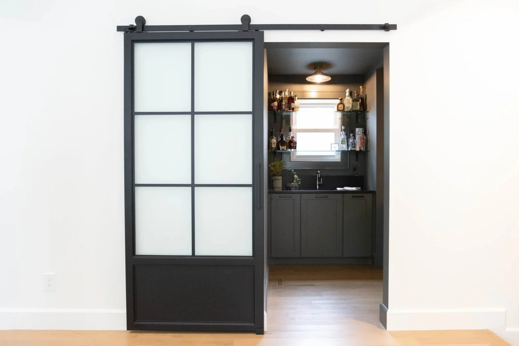 Neutral interior featuring a partially open sliding door revealing a moody scullery and wet bar with dim lighting, a sink, shelves with bottles, and a window.