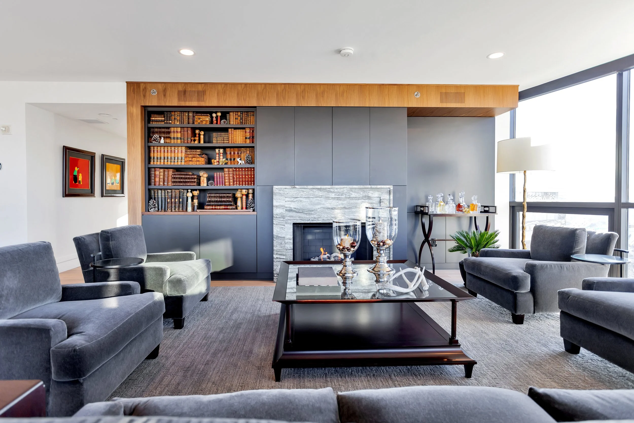Modern living room with grey sofas, a glass coffee table, and a stunning stone fireplace. Bookshelves and colorful art decorate the walls. Bright and cozy ambiance.