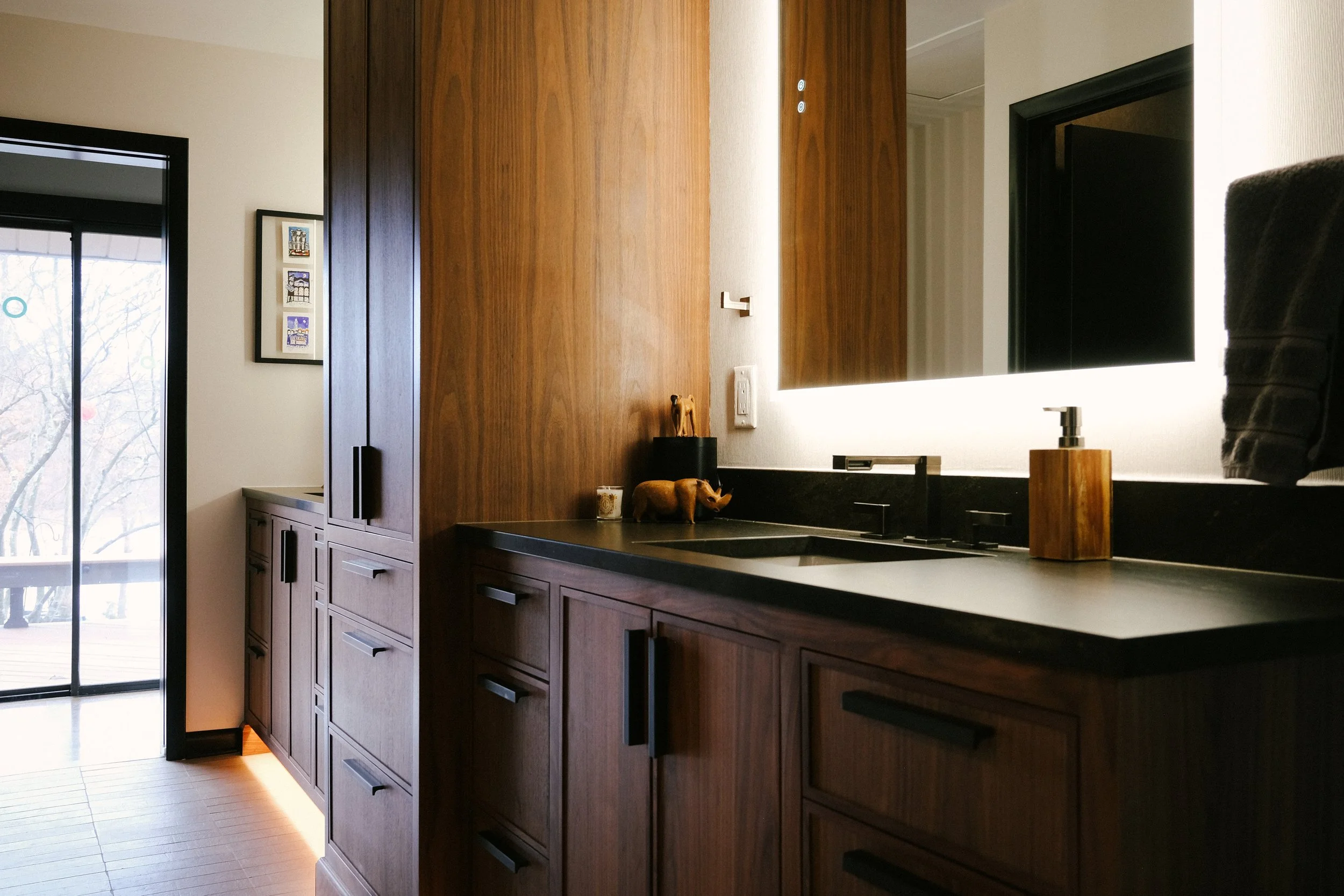 Modern bathroom with custom Walnut cabinets, Cambria Remington Brass countertop, and LED illuminated mirror. Sunlight filters through a window, creating a warm, inviting atmosphere.
