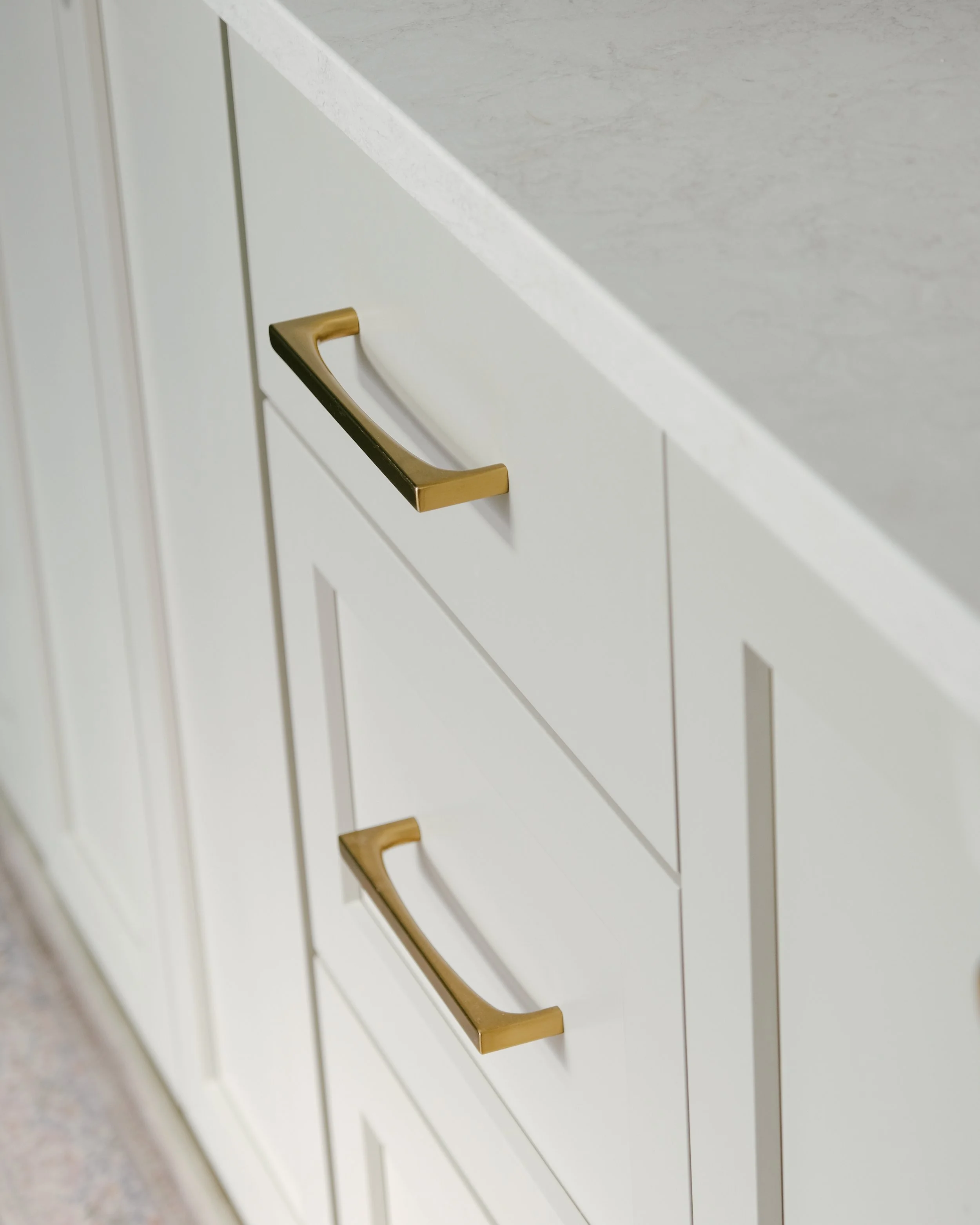 White custom bathroom cabinets with sleek honey bronze handles beneath a light quartz countertop. The design conveys elegance and minimalism.