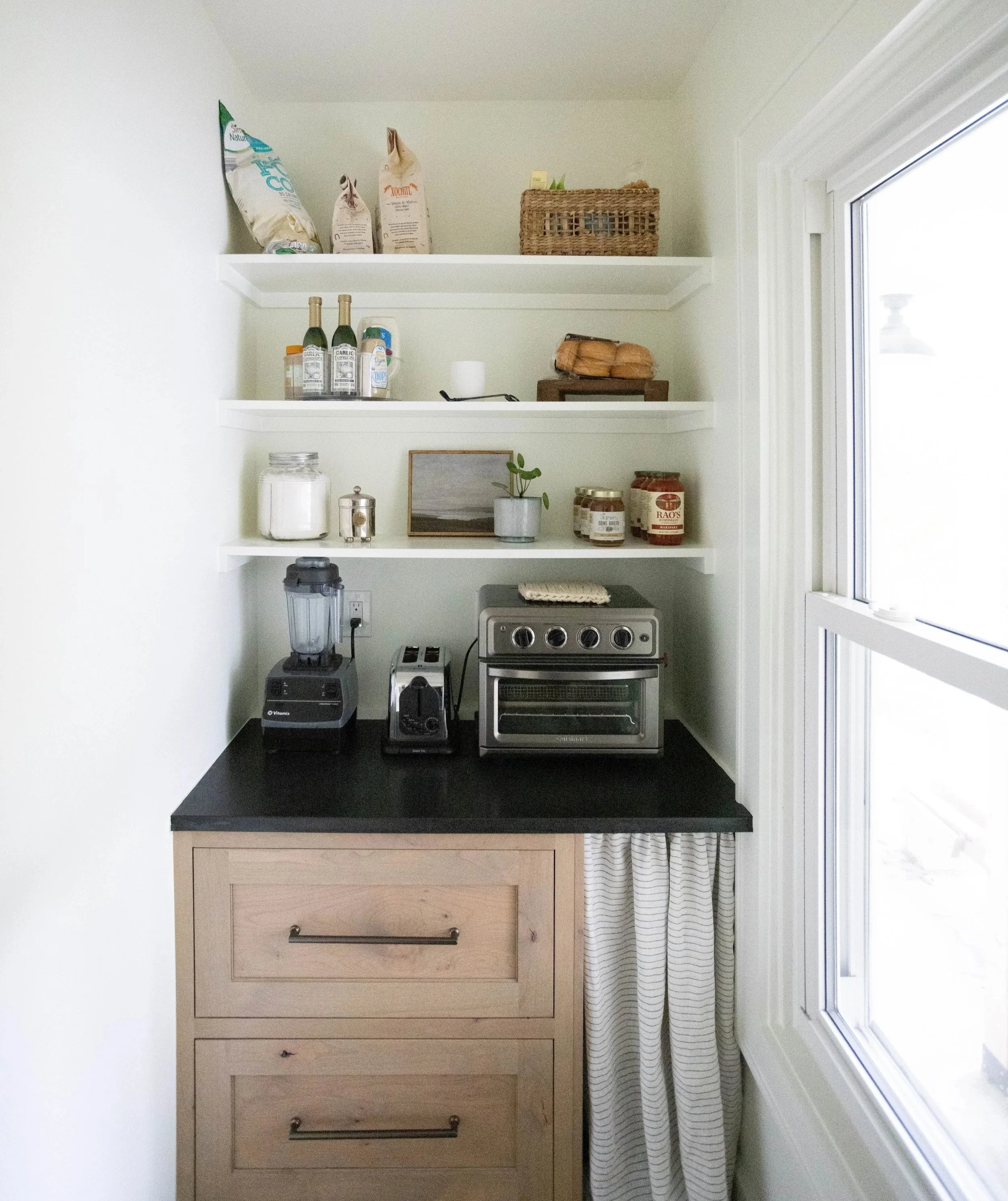 Compact kitchen pantry with open shelves holding jars, condiment bottles, a plant, and dry goods. A black quartz countertop is paired with natural wood lower cabinets and a linen curtain.