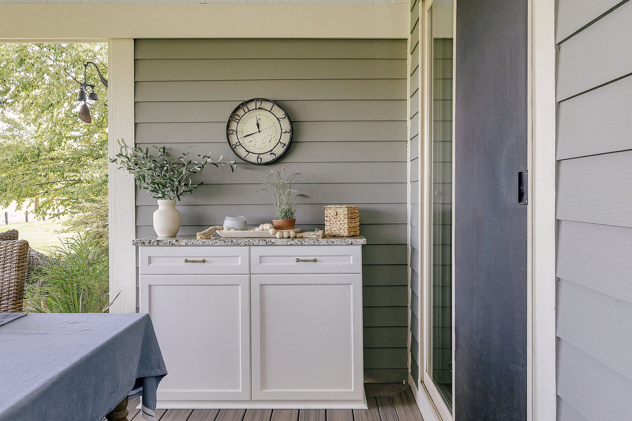 Outdoor patio with white custom cabinetry topped with plants, a candle, and wicker baskets. A clock hangs on the gray siding, conveying a serene setting.