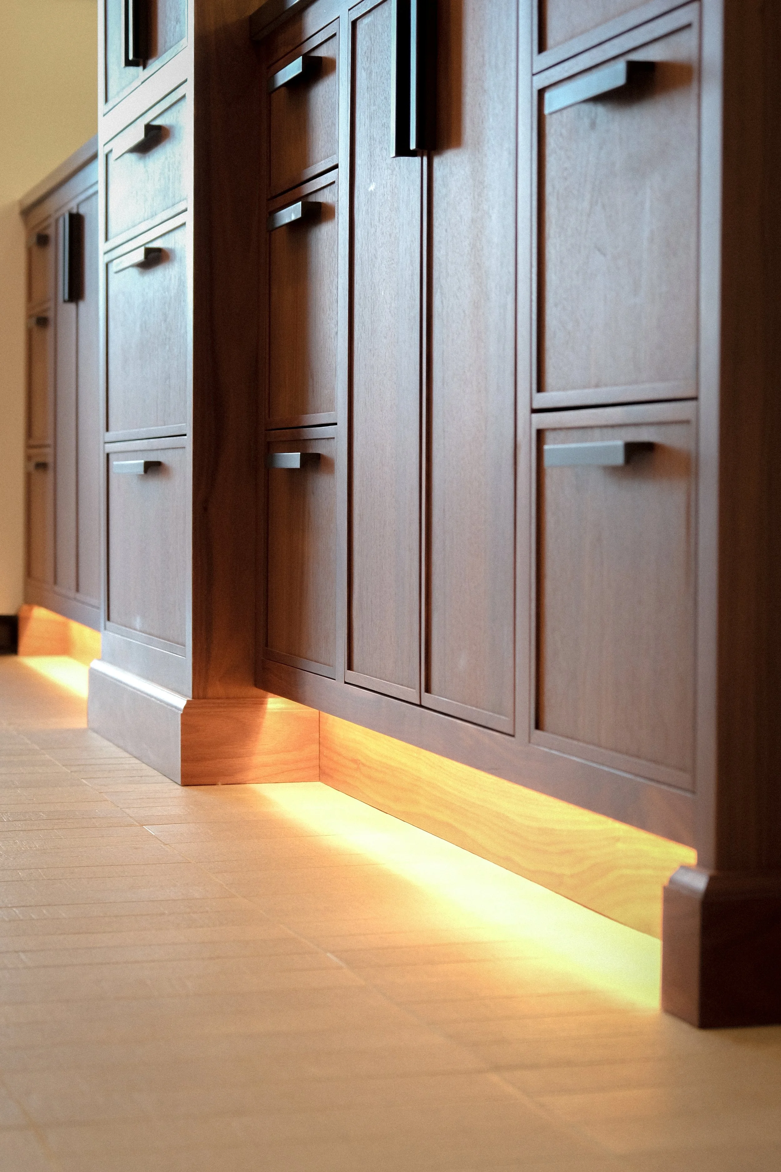 Custom Walnut vanity cabinets with matte black hardware line a modern bathroom, softly illuminated by warm under-cabinet lighting.