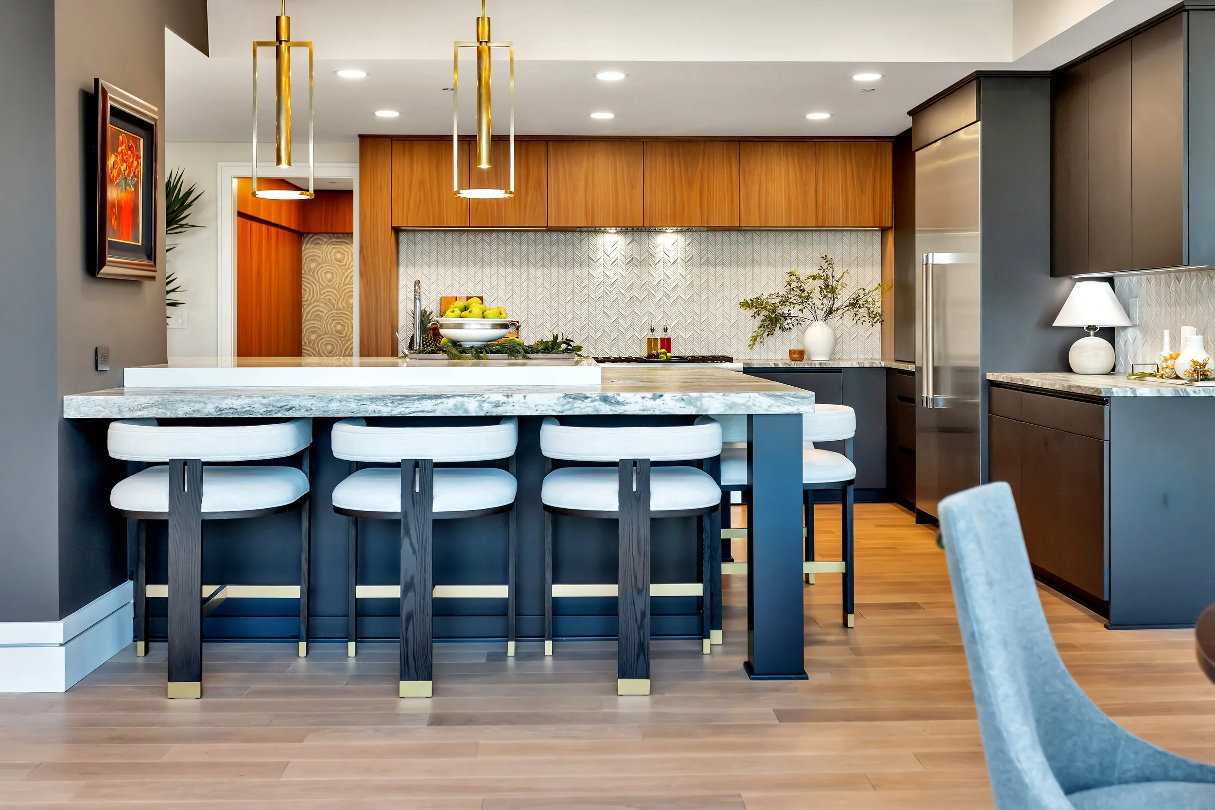 Contemporary kitchen with dark, moody custom cabinetry, accented by a large wooden range hood and quartz island with five upholstered stools for gathering. Neutral tones and elegant decor create a sophisticated ambiance.