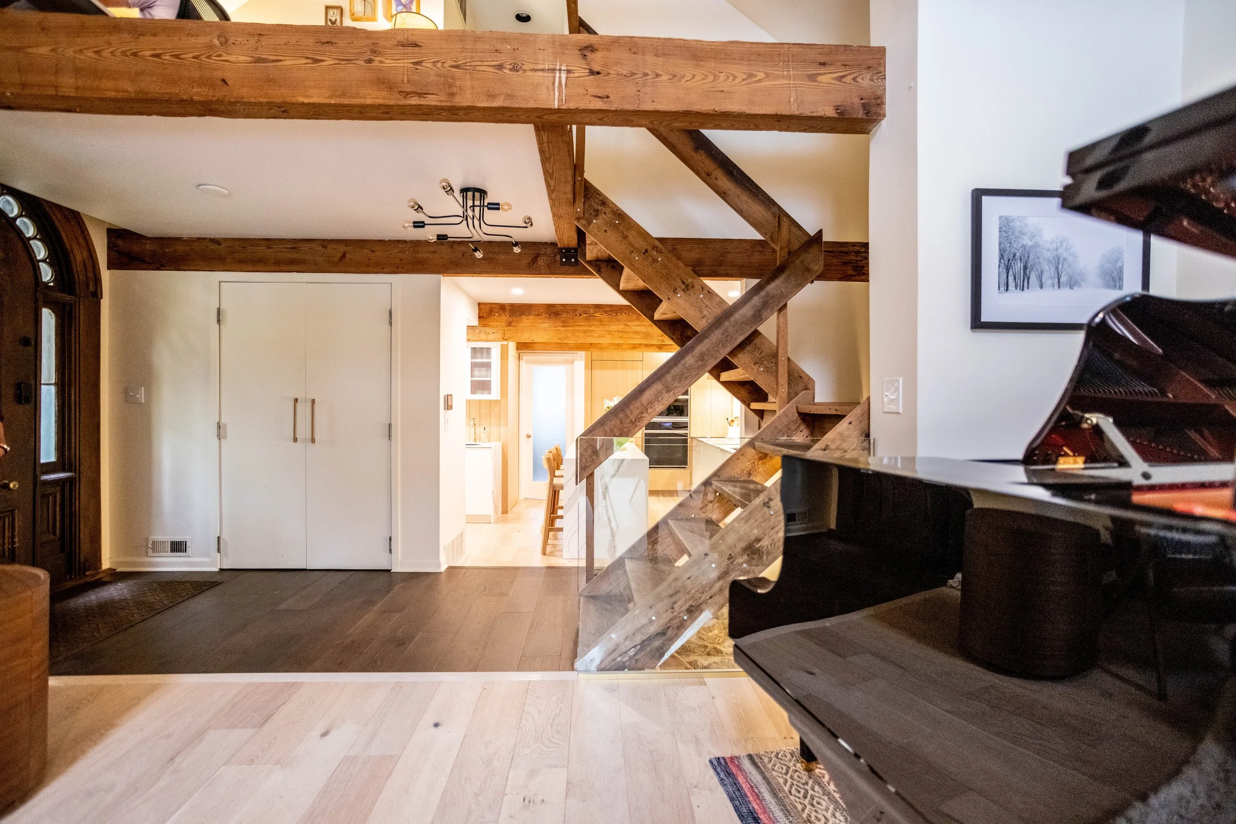Spacious rustic, minimalist interior with a wooden staircase, exposed beams and railing, dark and light wood floors, a grand piano, and cozy lighting, creating a warm ambiance.
