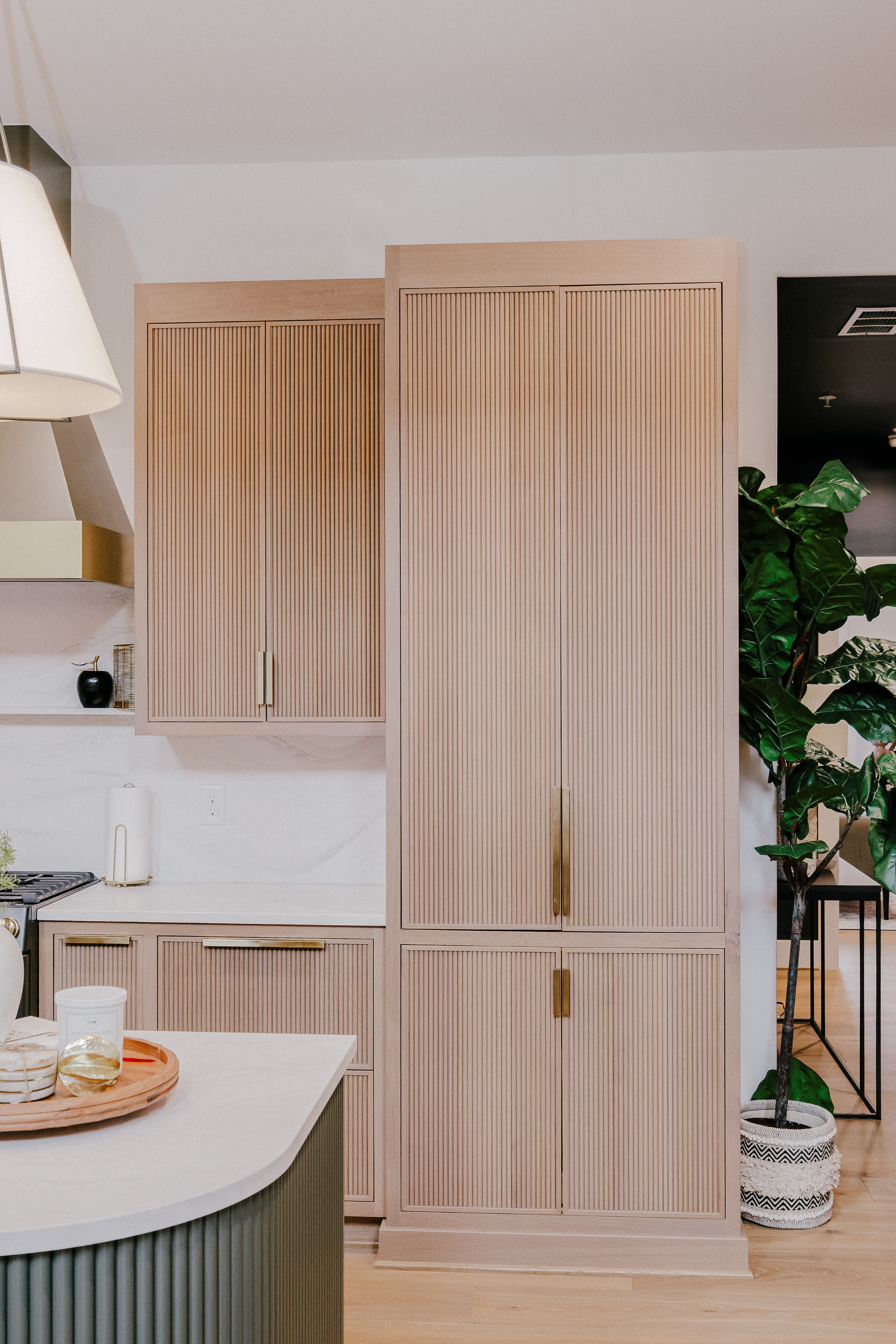 Modern kitchen features light wood reeded cabinets, a white quartz countertop, and a large green potted plant, creating a clean, inviting atmosphere.