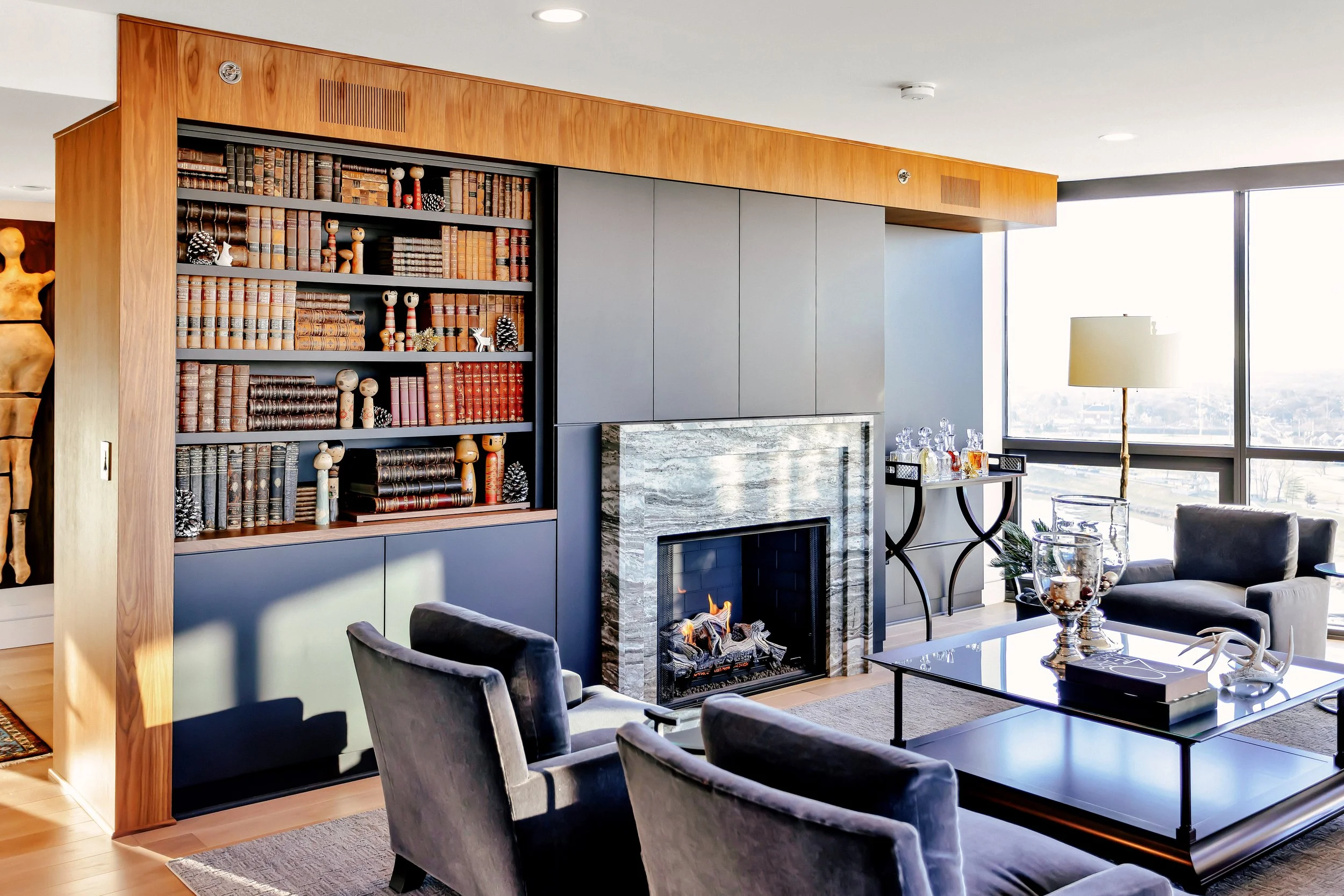 Modern living room with grey sofas, a glass coffee table, and a stunning stone fireplace. Bookshelves and colorful art decorate the walls. Bright and cozy ambiance.