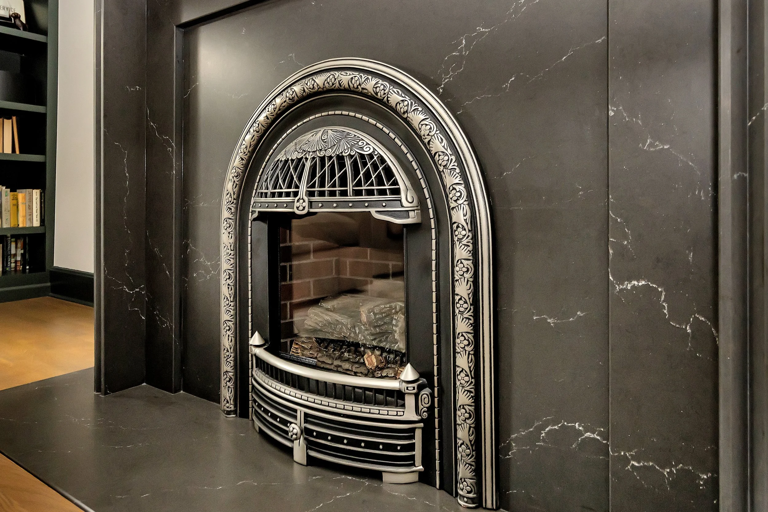 Elegant black fireplace with intricate silver detailing, ornate arch, and a cozy ambiance. Bookshelf in background adds a classic touch.