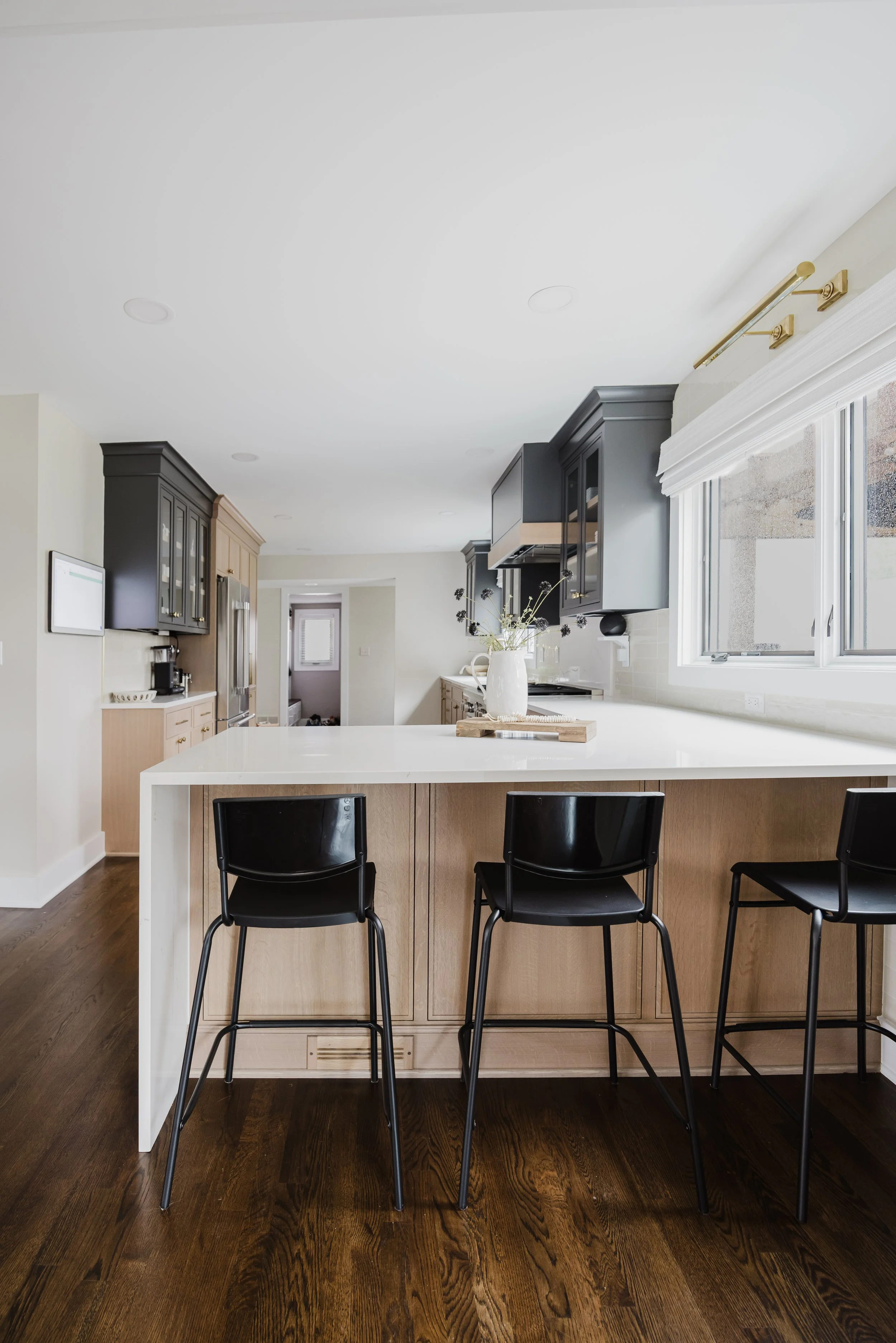Modern, neutral kitchen with a large white quartz island, three black barstools, black custom upper cabinets and natural White Oak cabinets, and dark wood flooring. Bright, airy atmosphere with natural light.