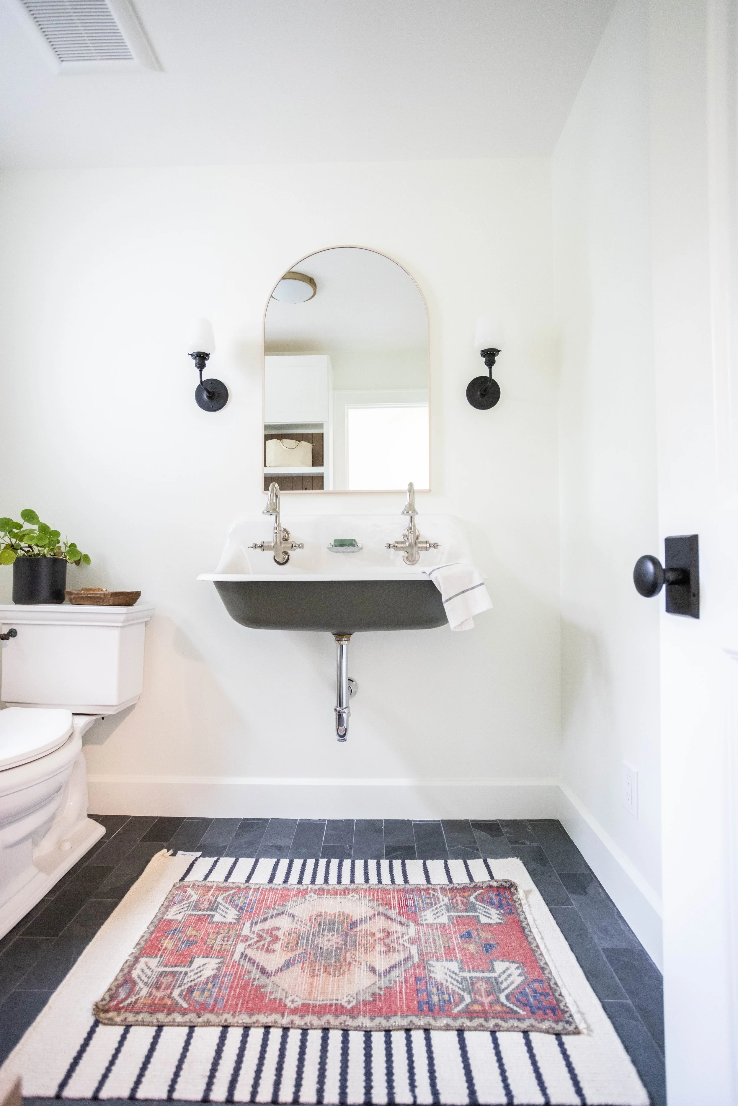Minimalist bathroom with a vintage-inspired dual-faucet sink, arched mirror, and wall-mounted sconces. A green plant sits on the toilet tank beside a woven basket.