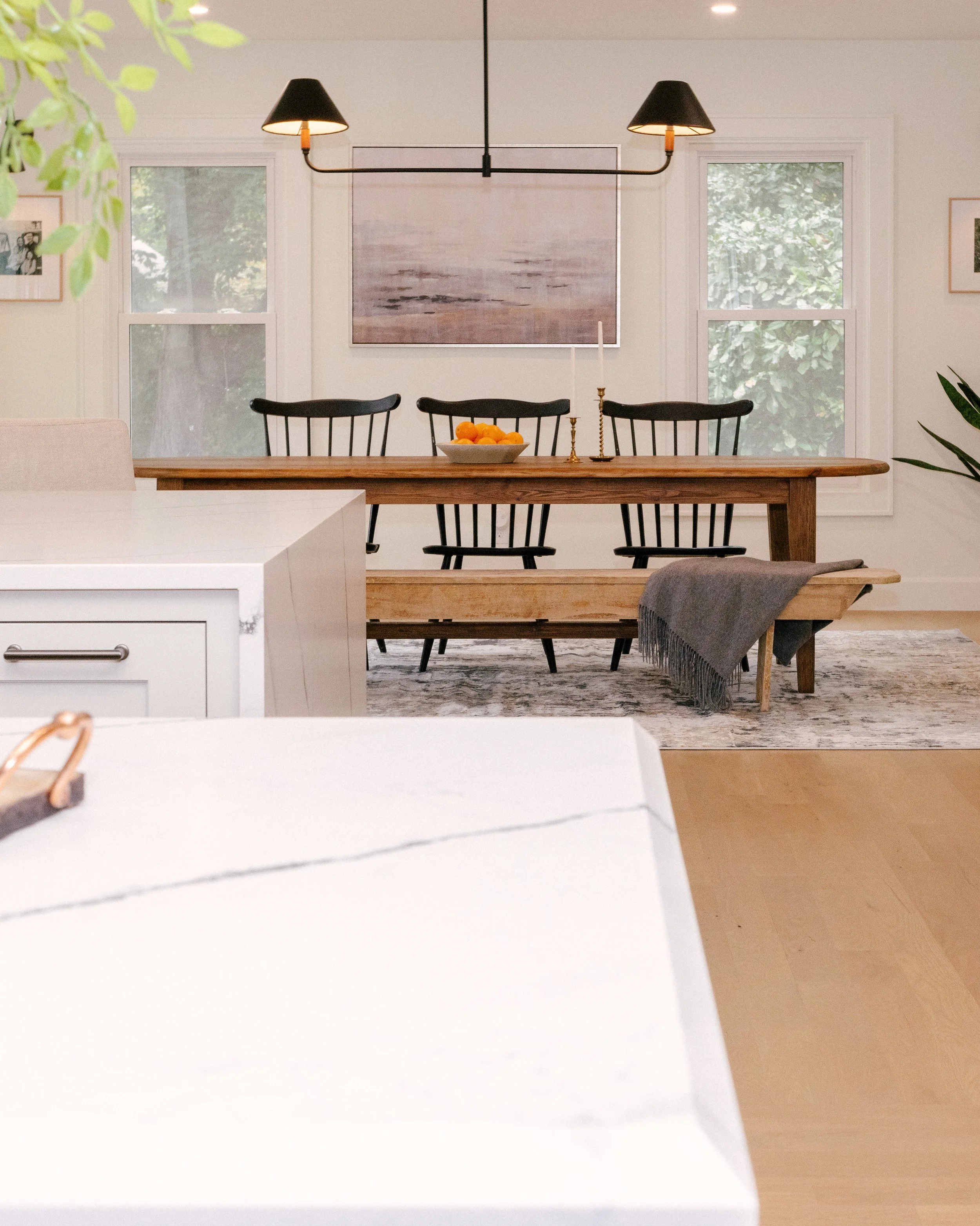Sleek, earthy dining area with a wooden table, black chairs, and a bench draped with a gray blanket. A minimalistic lighting fixture sits above.
