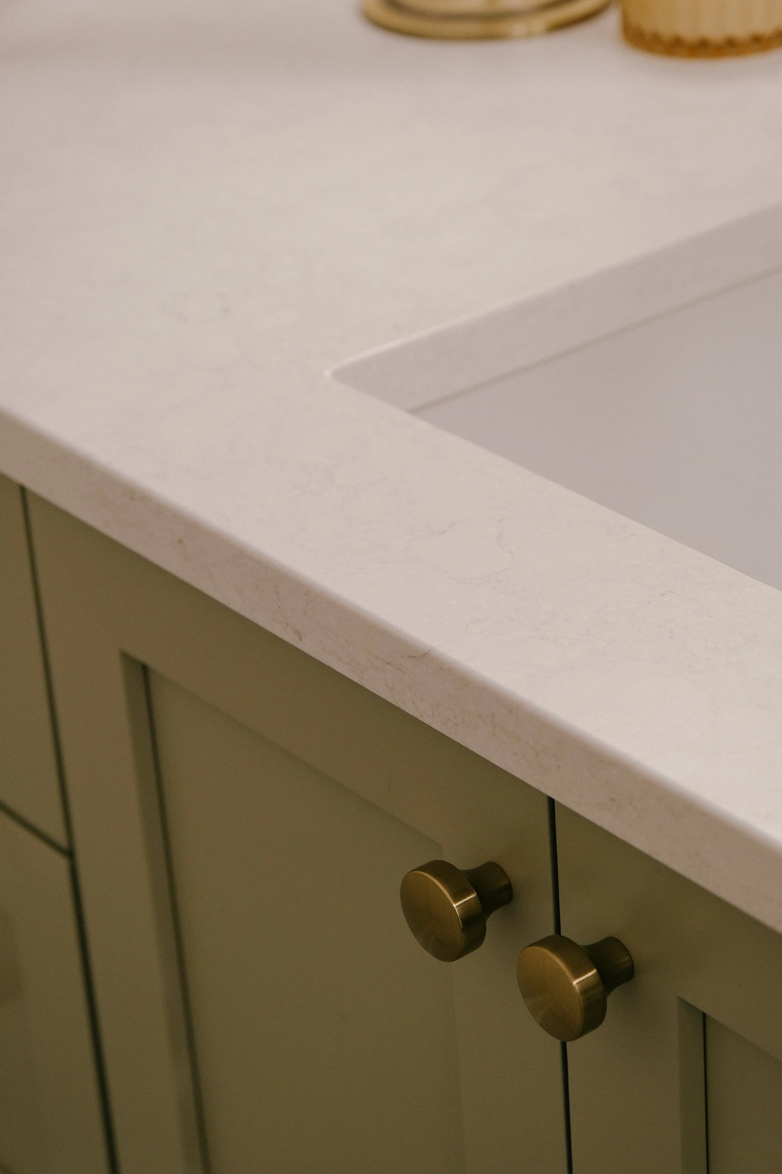 Close-up of a countertop and cabinet corner. The countertop is subtly veined quartz, and the cabinet features slate green doors with gold hardware, providing an elegant, modern look.