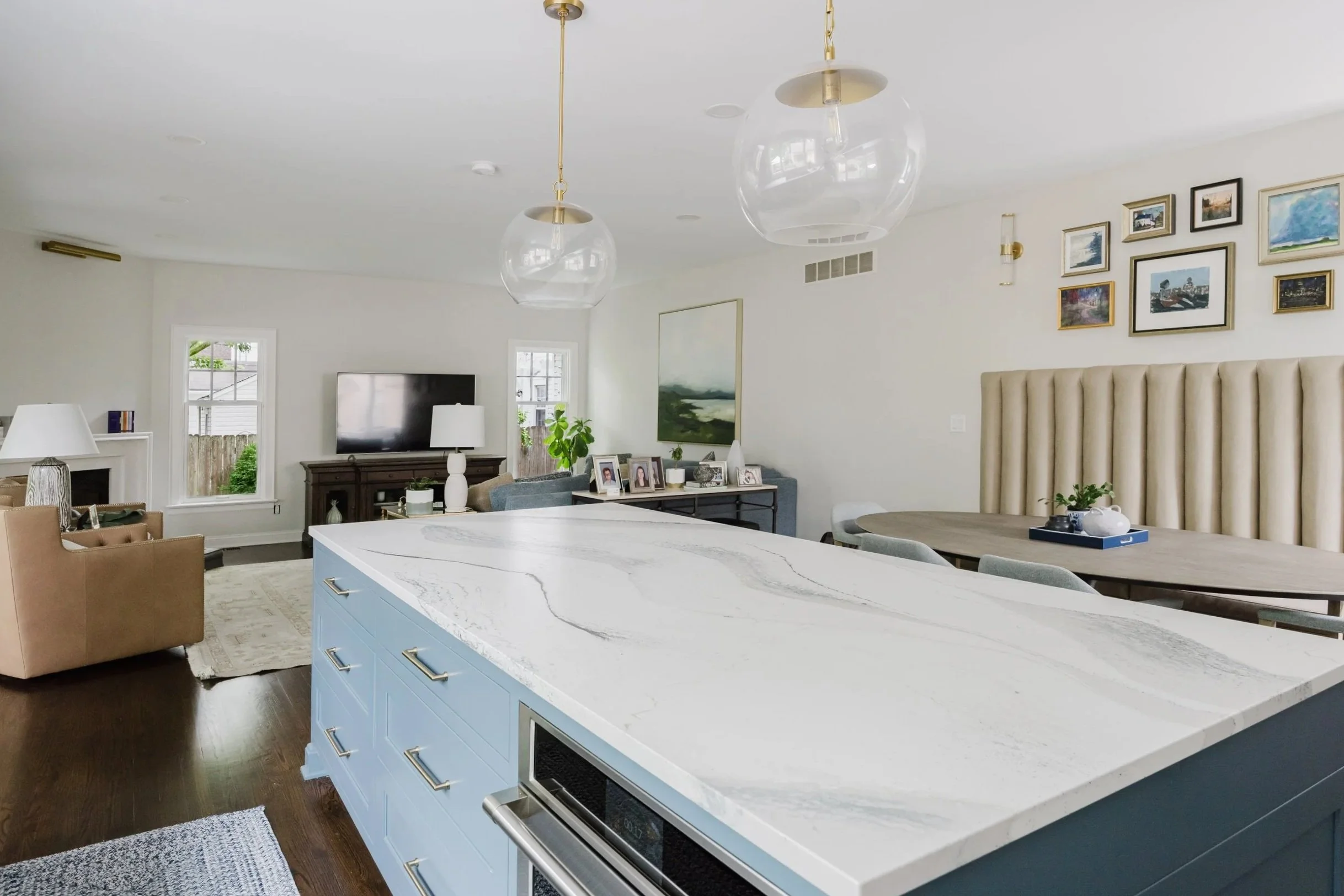 Modern living room and kitchen with blue island, Cambria quartz countertop, pendant lights. In a living just beyond the kitchen sits a TV, soft, minimalist seating, a stone fireplace and framed artwork on walls. Warm and inviting.