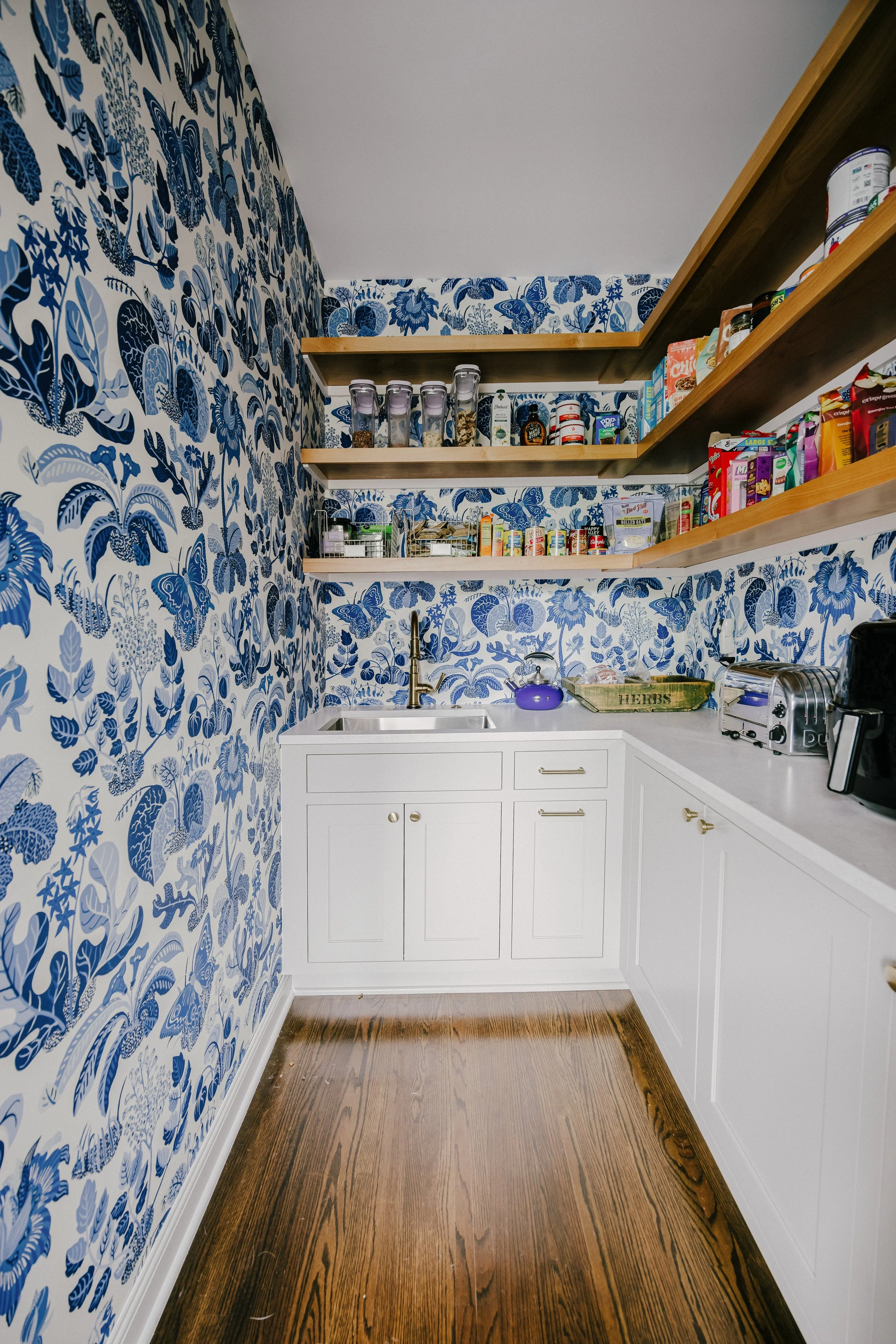 Quaint pantry with blue floral wallpaper, wooden shelves stocked with jars and cans, a small Elkay sink, gold Delta faucet, and a blue kettle. Cozy and organized atmosphere.