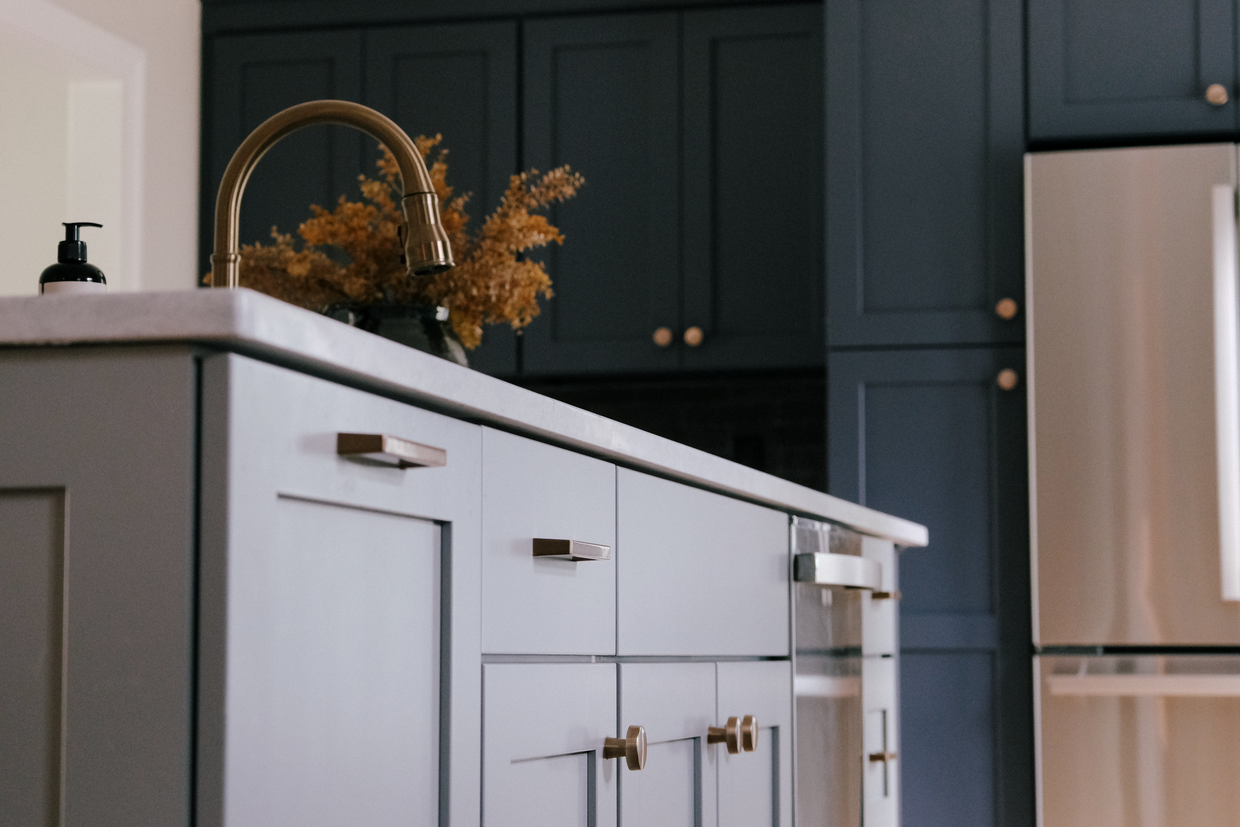 Modern, urban kitchen with grey and blue custom cabinetry, gold accents, and stainless steel appliances. A vase with dried flowers adds a warm touch.