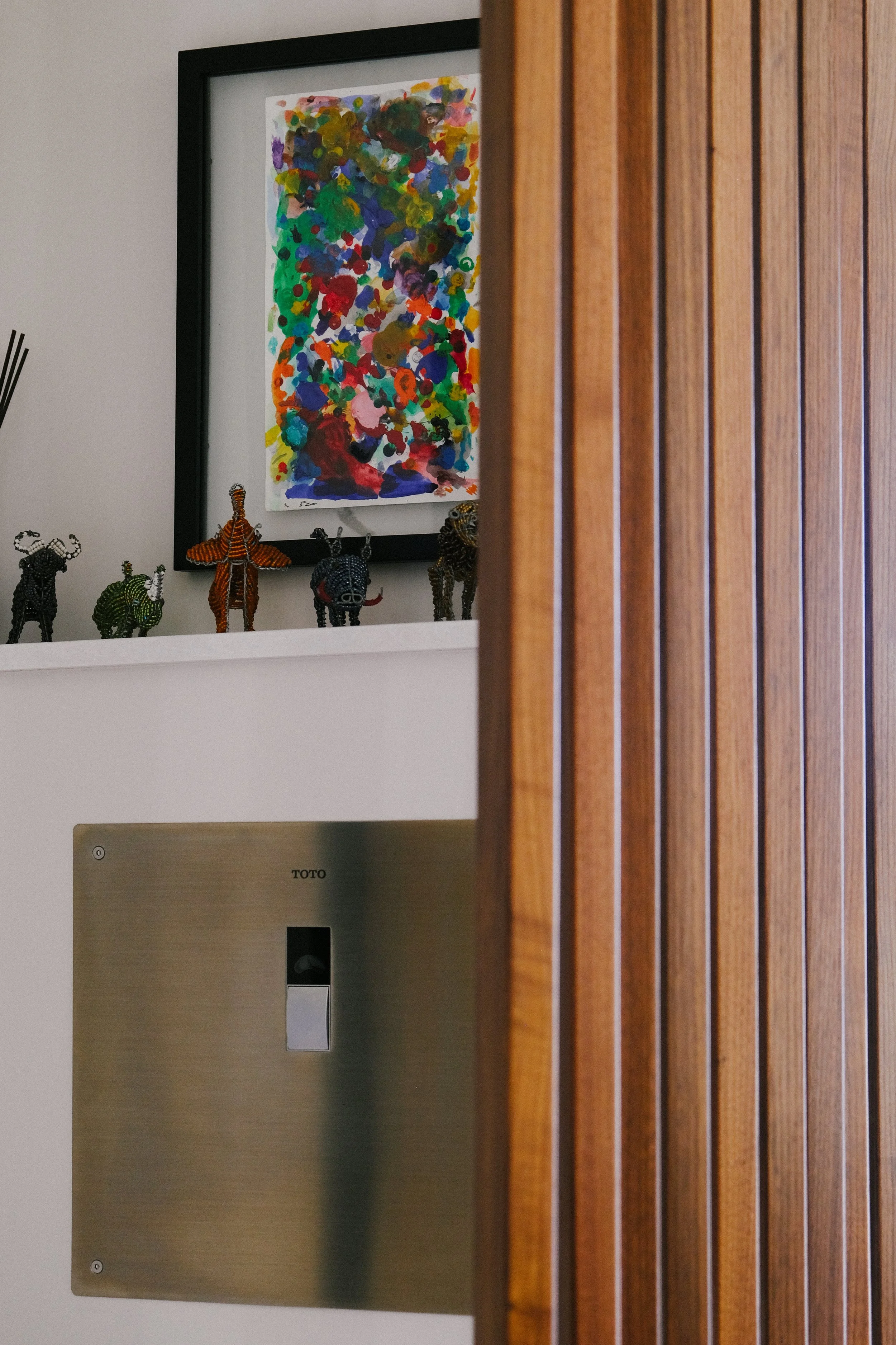 A colorful abstract painting in a black frame is on a shelf with beaded animal figurines. A reeded divider wall adds modernism and privacy to the urinal space.