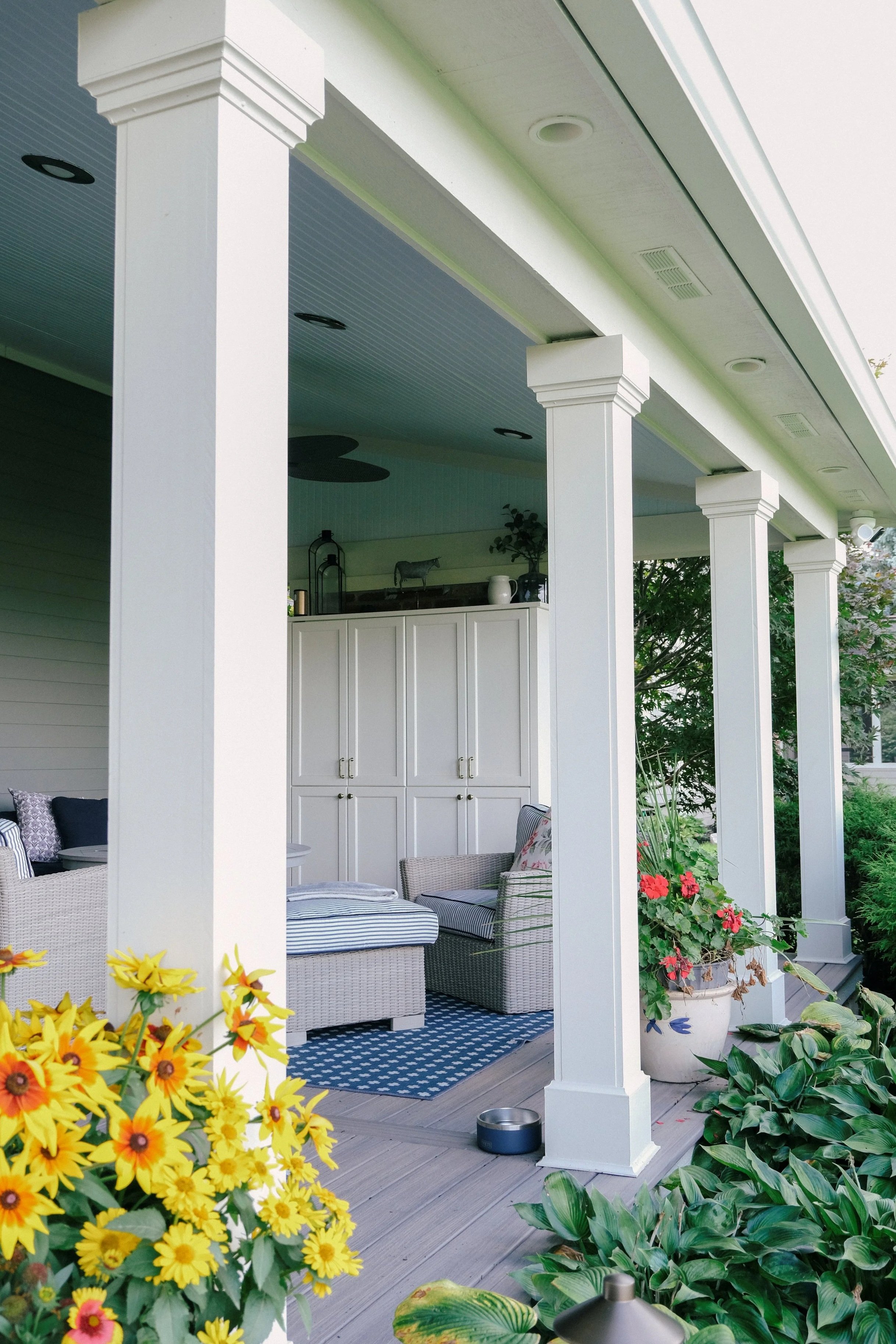 Covered porch with white columns and wicker furniture. Yellow flowers and lush greenery add a vibrant touch. Cozy, serene outdoor space.