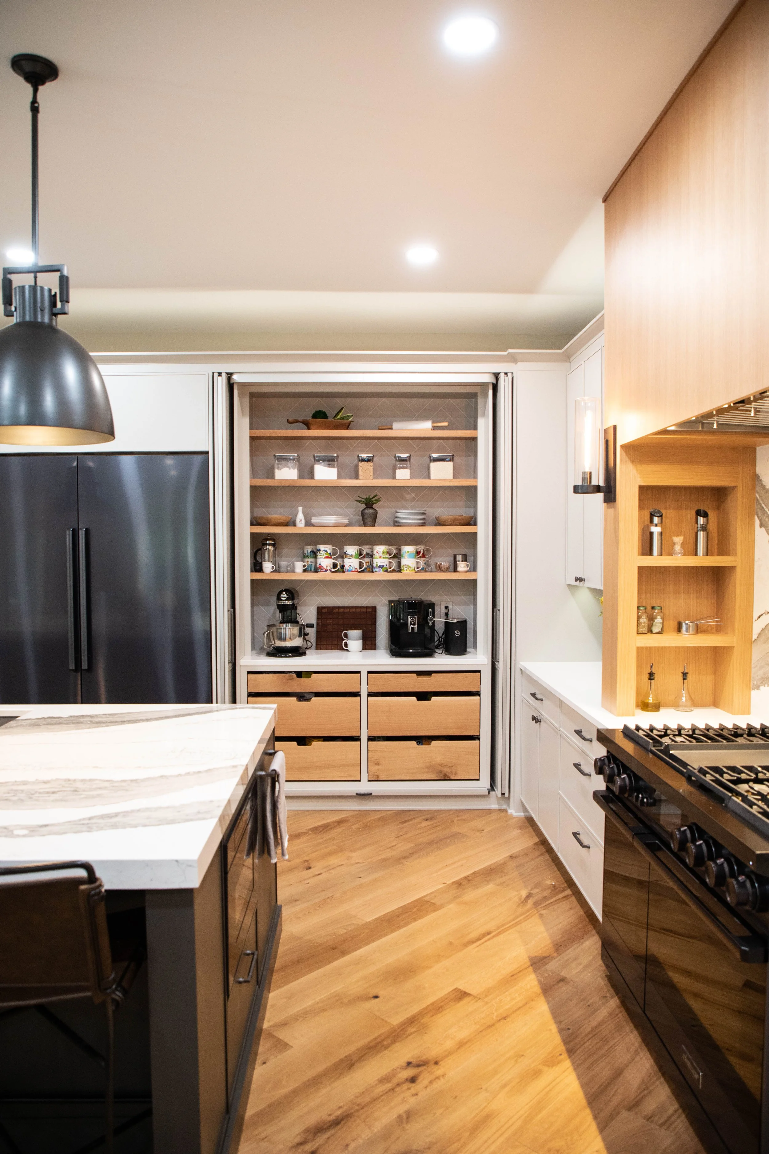 Pocket doors open to reveal an inset coffee bar in a modern kitchen. Next to the coffee bar is a black refrigerator.