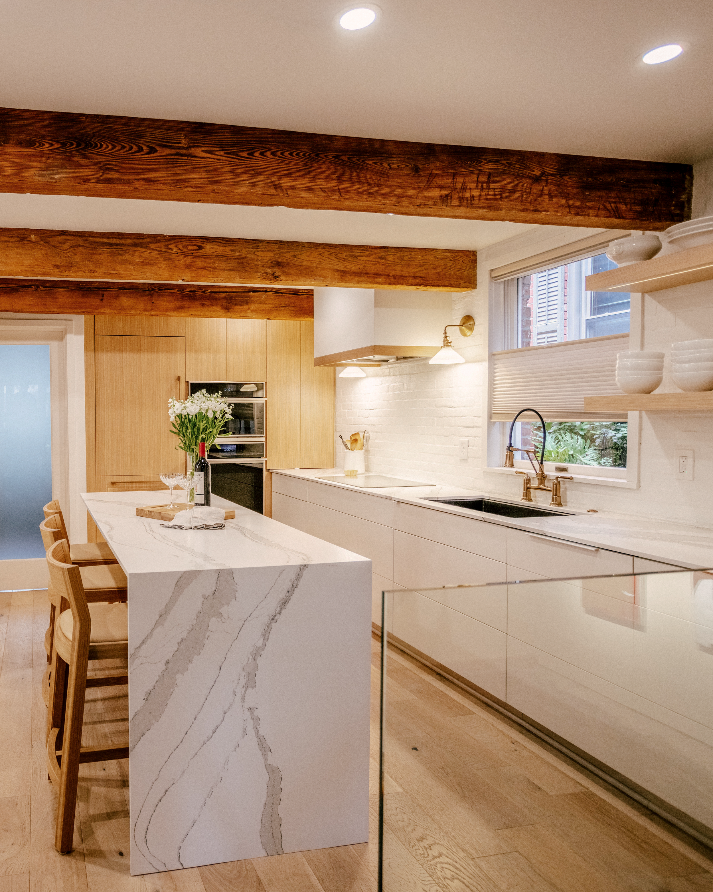 Scandinavian, minimalistic kitchen with exposed wood beams, quartz waterfall island, and wooden stools. A White Oak appliance wall, white painted brick backsplash, open wooden shelving, and a vase of flowers add warmth.