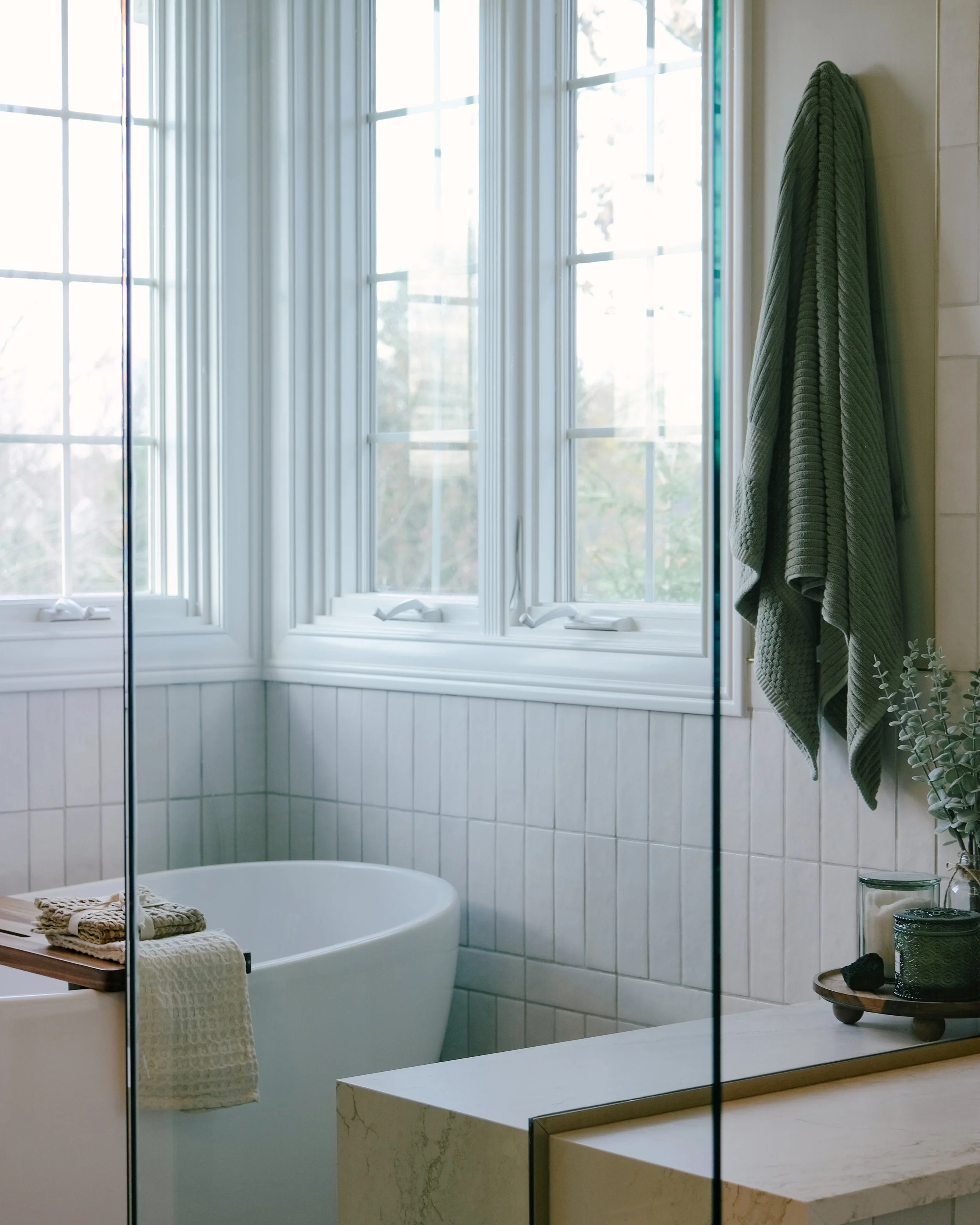 Bright bathroom with large windows, a white freestanding tub, and a green towel hanging. A waterfall shower bench holds candles and a small plant.