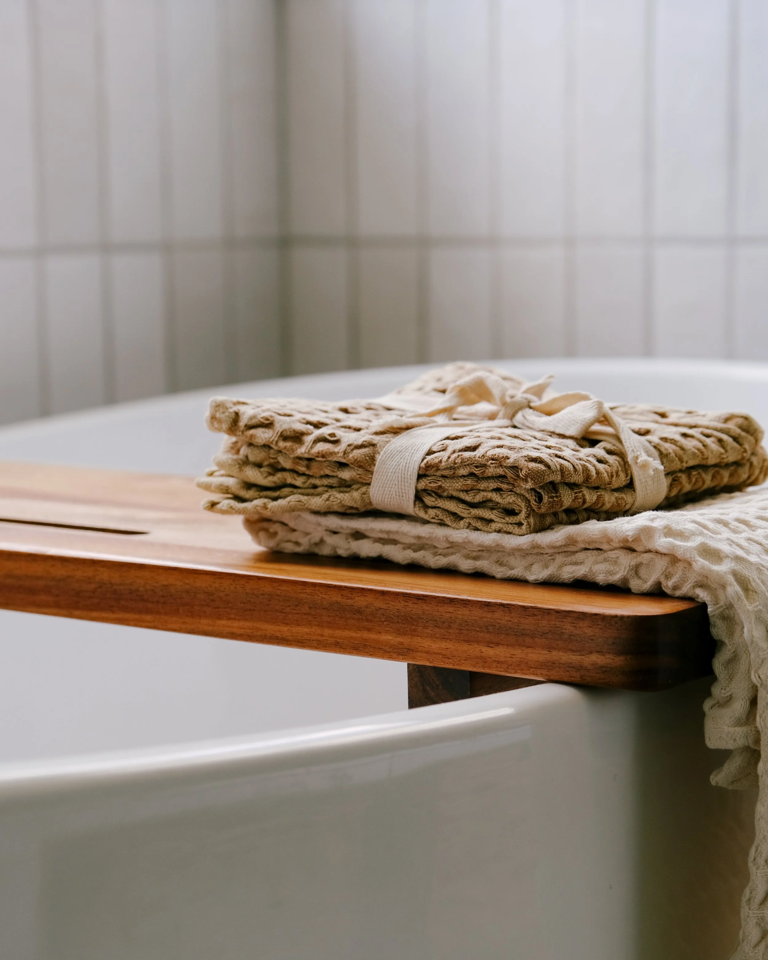 Folded beige towels neatly placed on a wooden bath tray over a white freestanding bathtub, with white tiled walls in the background, conveying a serene spa-like atmosphere.