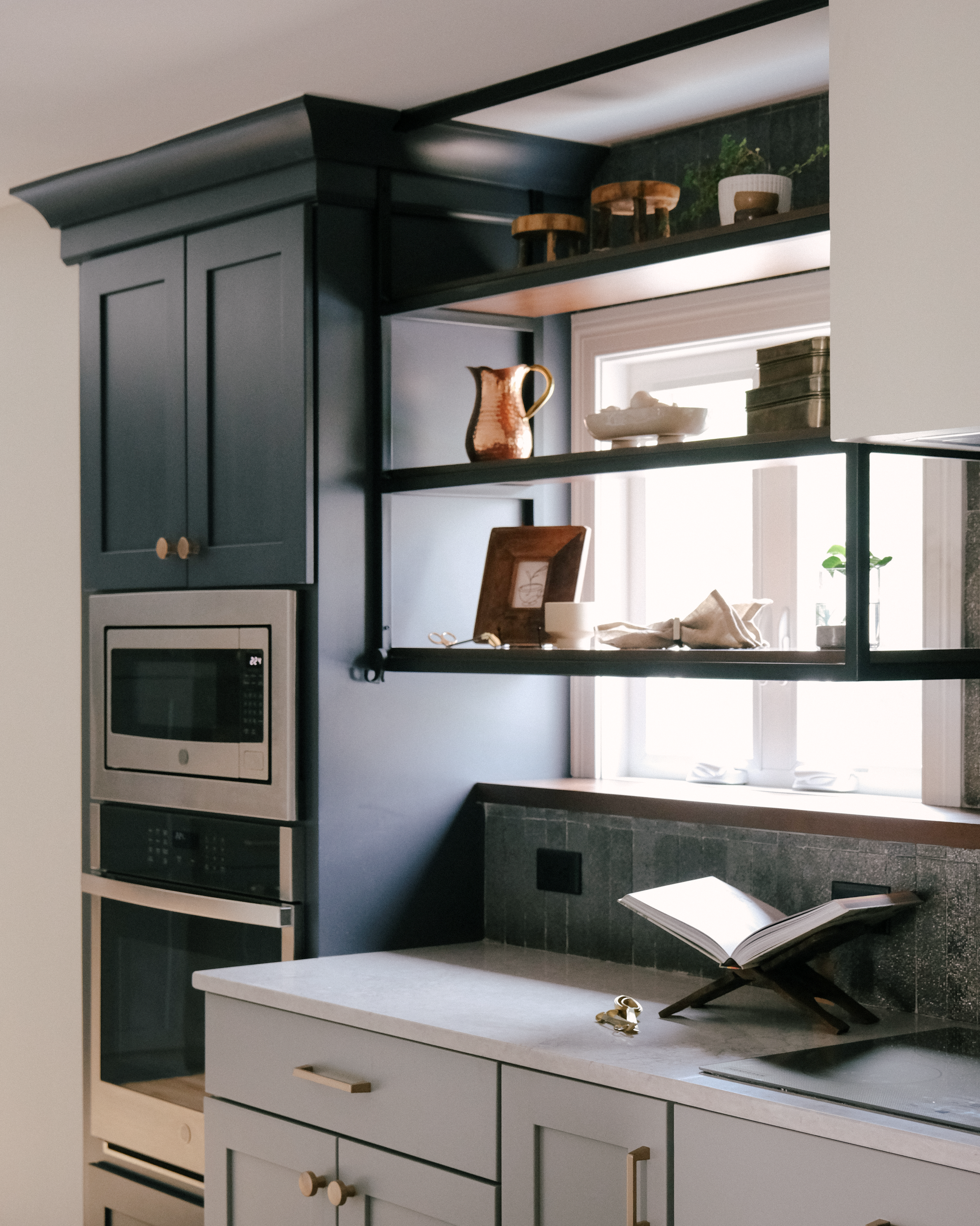 Modern kitchen corner with modern dark blue cabinets, built-in microwave and oven. Open industrial metal shelves display decor near a sunlit window.
