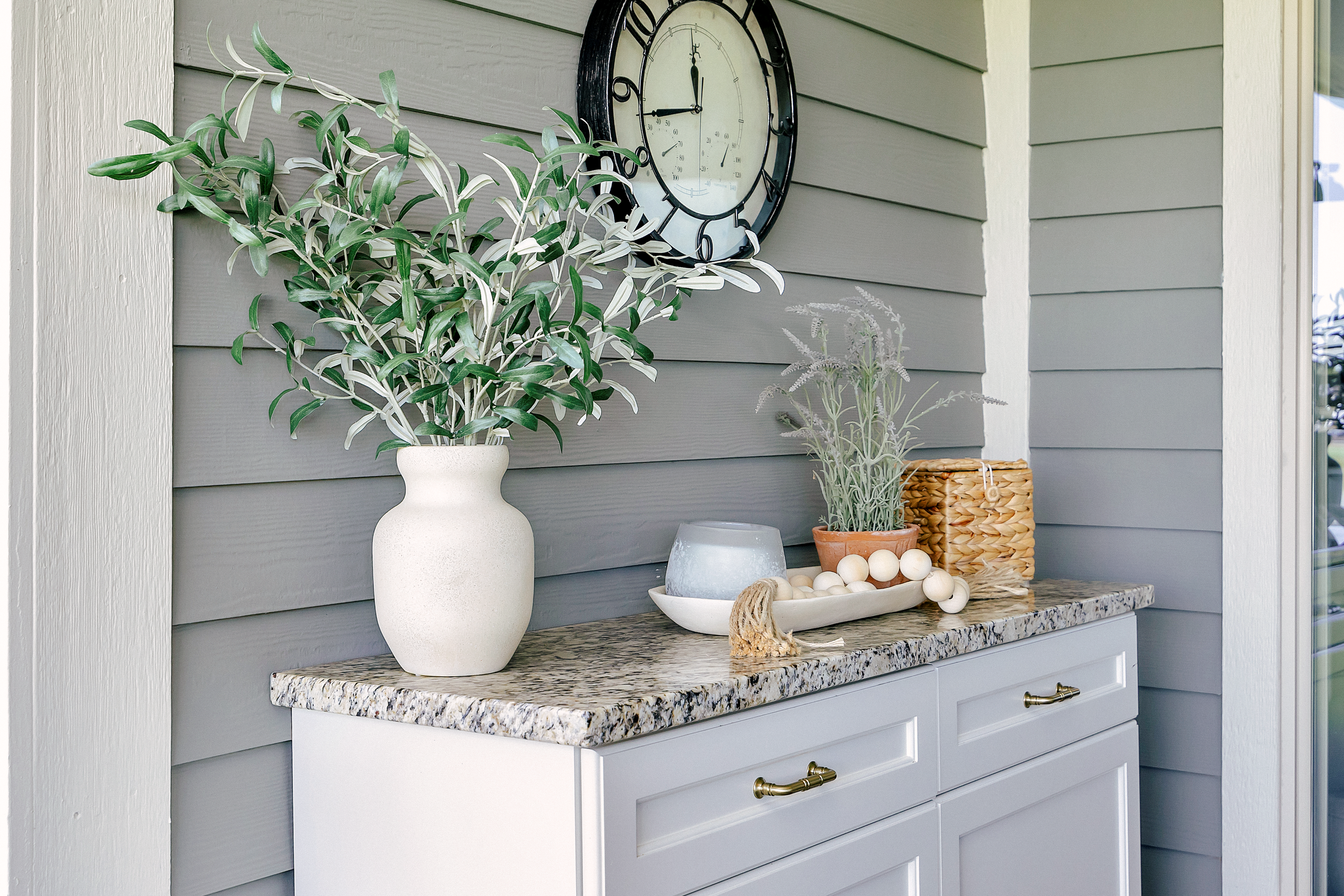 A custom outdoor cabinet with honey bronze hardware accents sits against chic grey siding. A weathered wall clock hangs above, evoking a rustic charm.