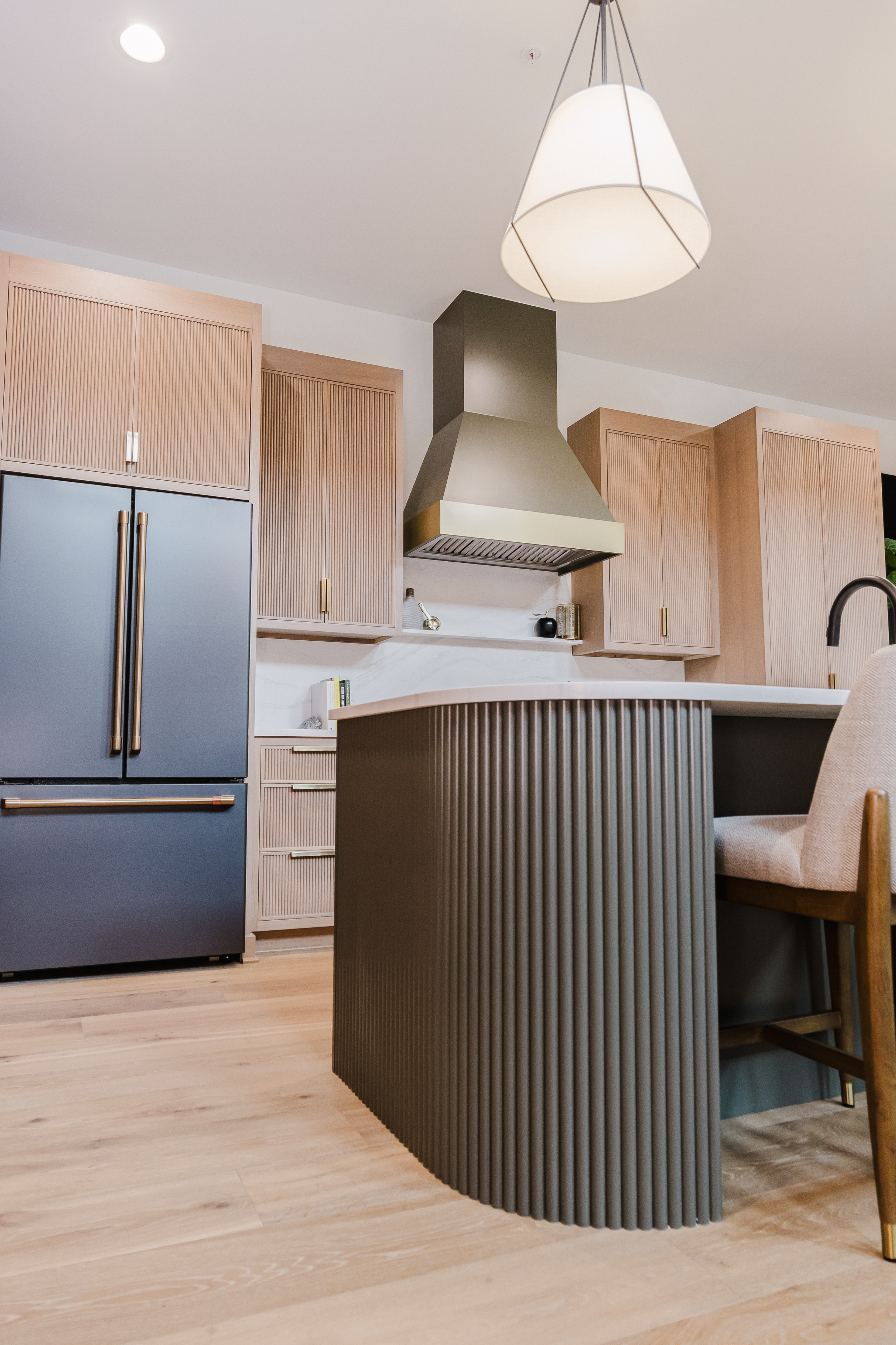 Modern kitchen with wooden reeded cabinets, a metallic range hood, and a sleek matte black fridge. A dark reeded island and chair sit below a large pendant light.
