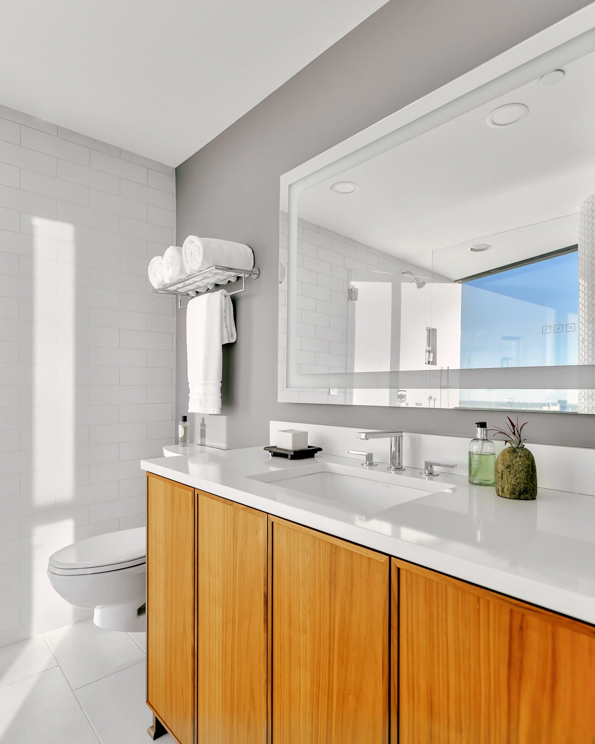 Modern bathroom with wooden vanity, large mirror, and white quartz countertop. Towels are neatly stacked on a wall rack. Bright and minimalistic ambiance.