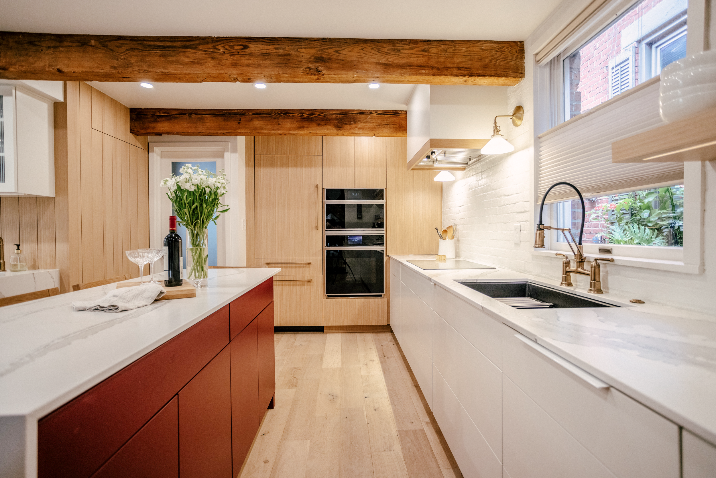 Scandinavian kitchen with exposed wooden beams, featuring red and white custom cabinetry. A White Oak appliance wall shows off an oven and paneled refrigerator under warm lighting.