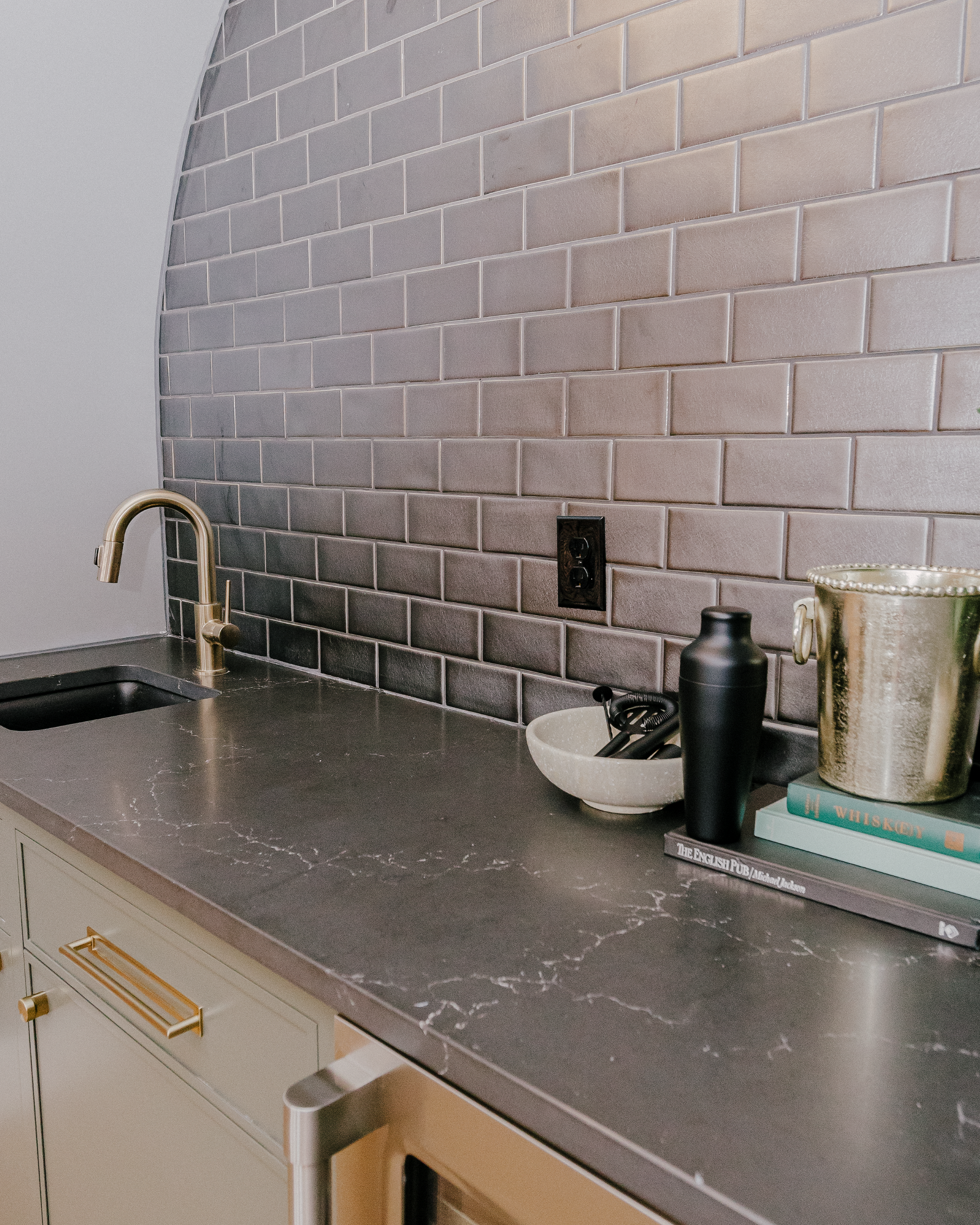 A beautiful, moody wet bar surrounded by a matte black tile backsplash. A bronze faucet and sink sit amidst black Cambria quartz countertops with subtle white veining.