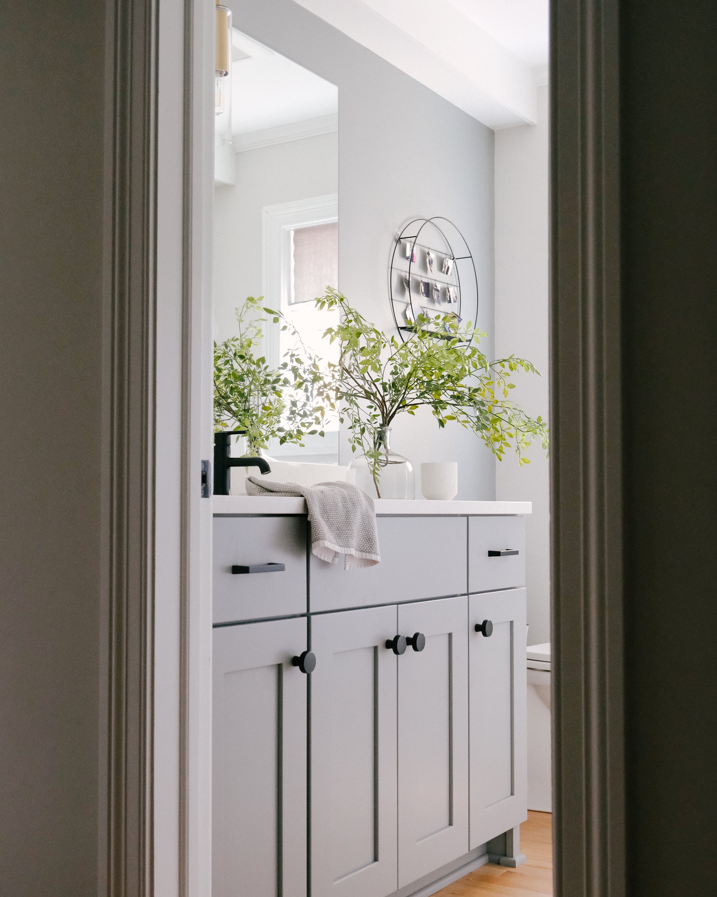 Gray bathroom vanity with black hardware and matte black faucet, topped with bright greenery in a vase. A soft towel hangs over the edge, under soft natural light.