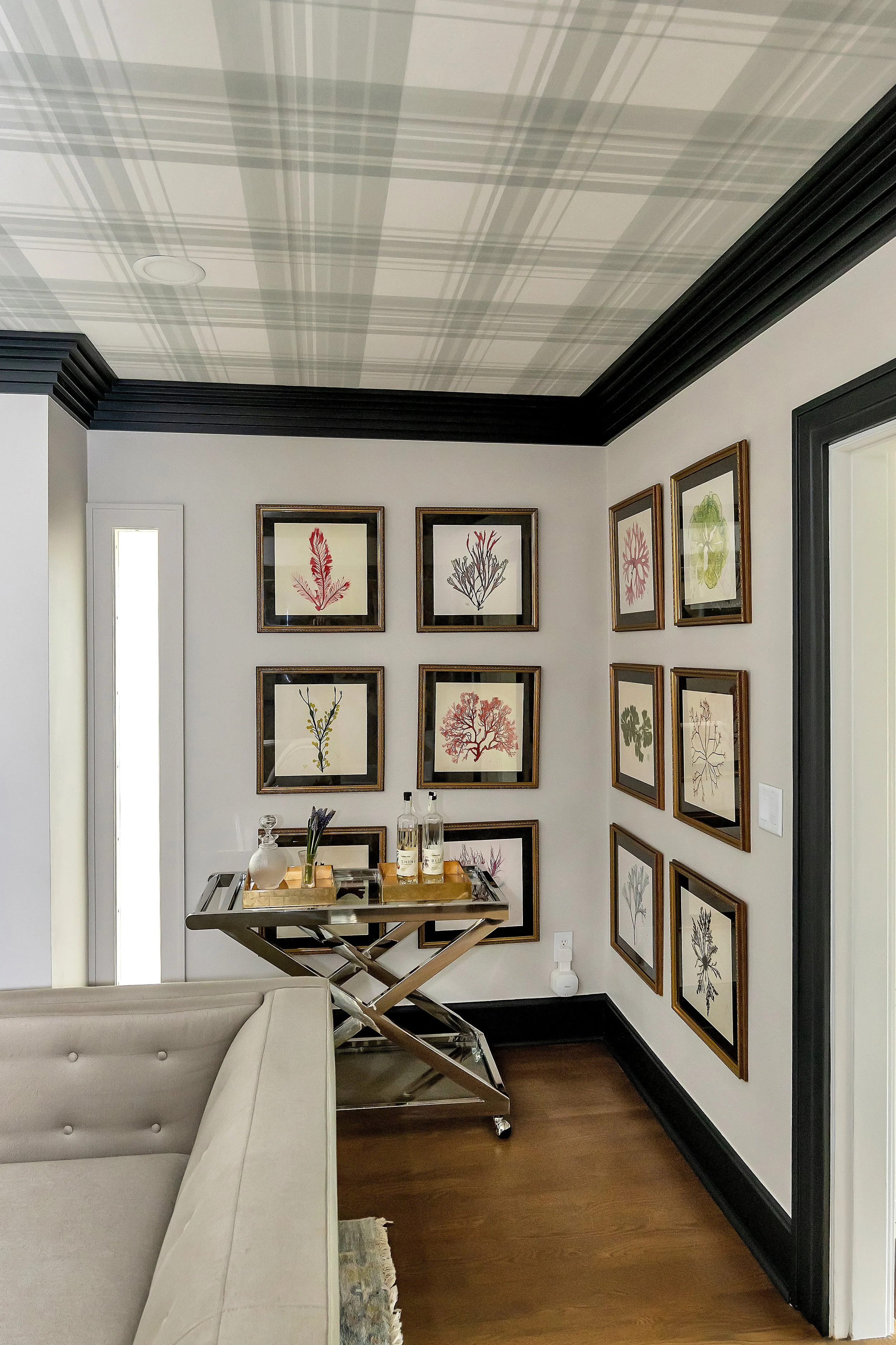 A stylish corner with framed botanical art on grey walls, a plaid wallpapered ceiling, and a sleek bar cart. The tone is elegant and inviting.