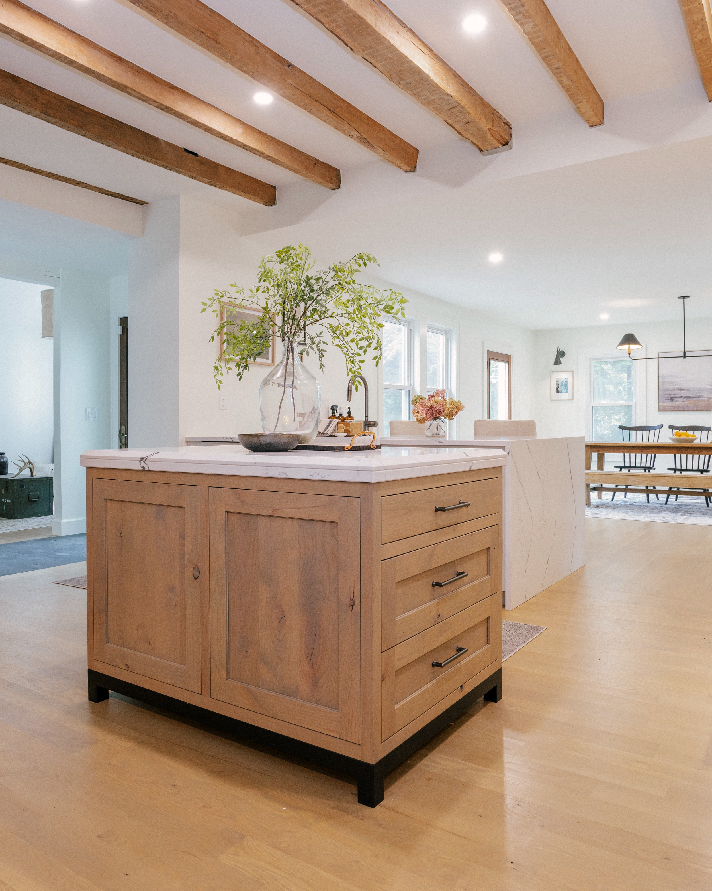 A modern kitchen with wooden beams. A central island features a natural, rustic wood finish, topped with a vase of greenery. A second island in the background features a Cambria quartz waterfall top.