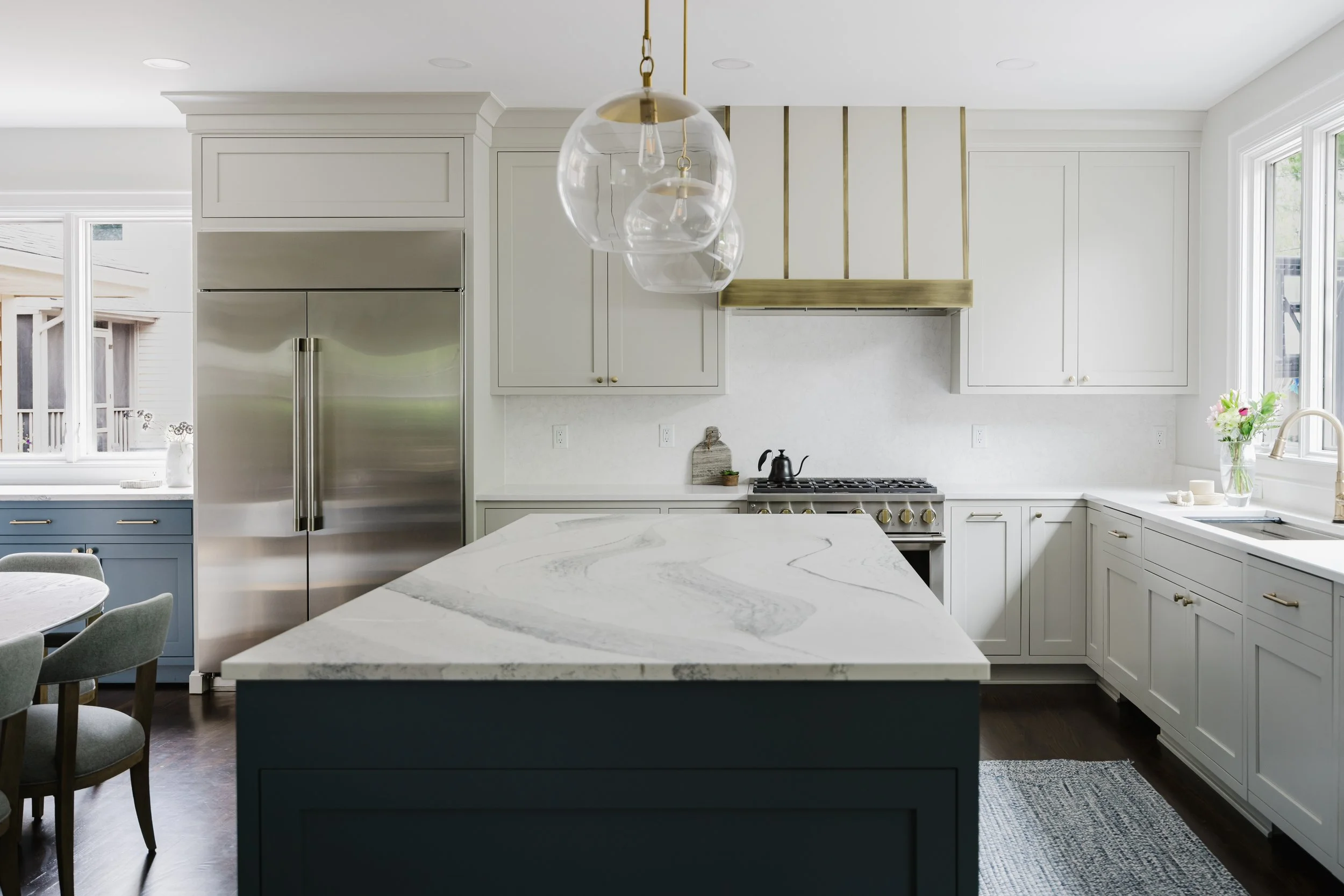 Modern kitchen with a ten-foot blue island, white custom cabinetry, and hardwood floors. Gold-accented pendant lights hang above. Dining area has a cushioned bench and framed art.