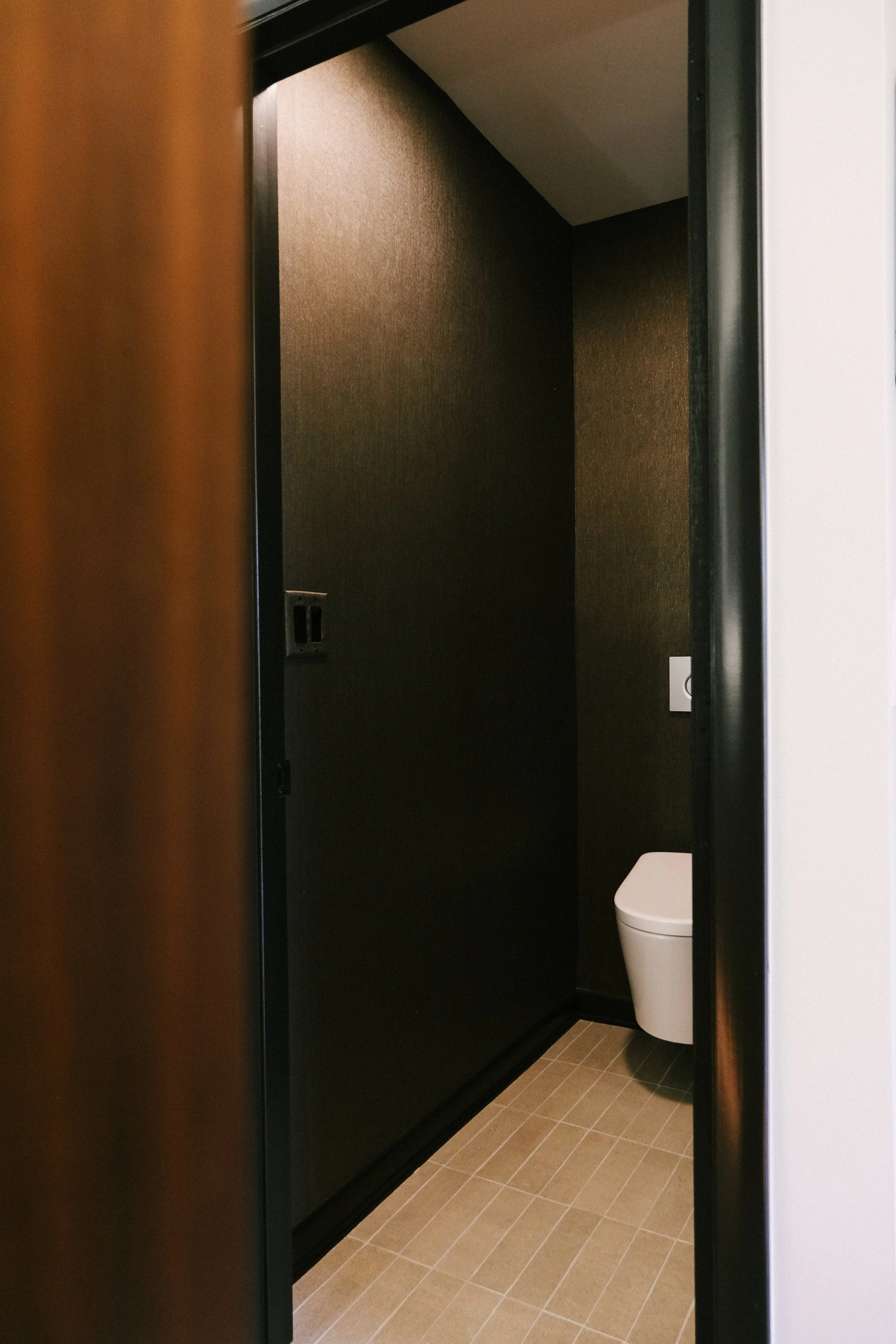 Moody bathroom with black, textured walls and beige tiled floor viewed through a partially open door. The white toilet adds a modern touch.