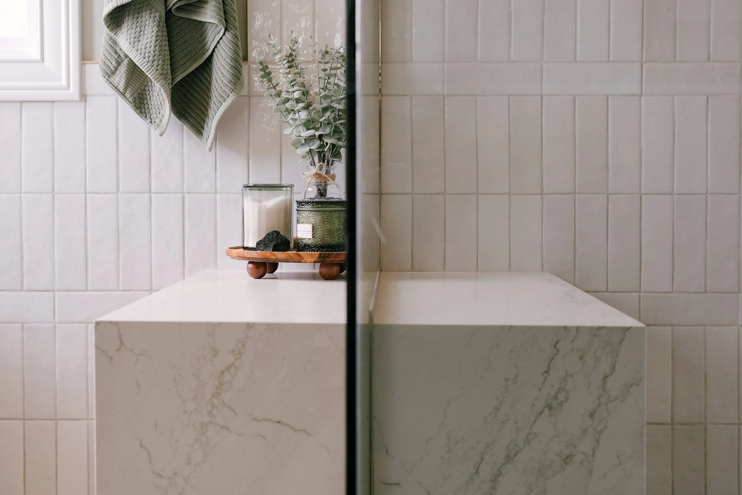 A gorgeous pass-through Cambria Everleigh waterfall shower bench holds a wooden tray, candles and eucalyptus against a white tiled shower. Minimalist and serene.
