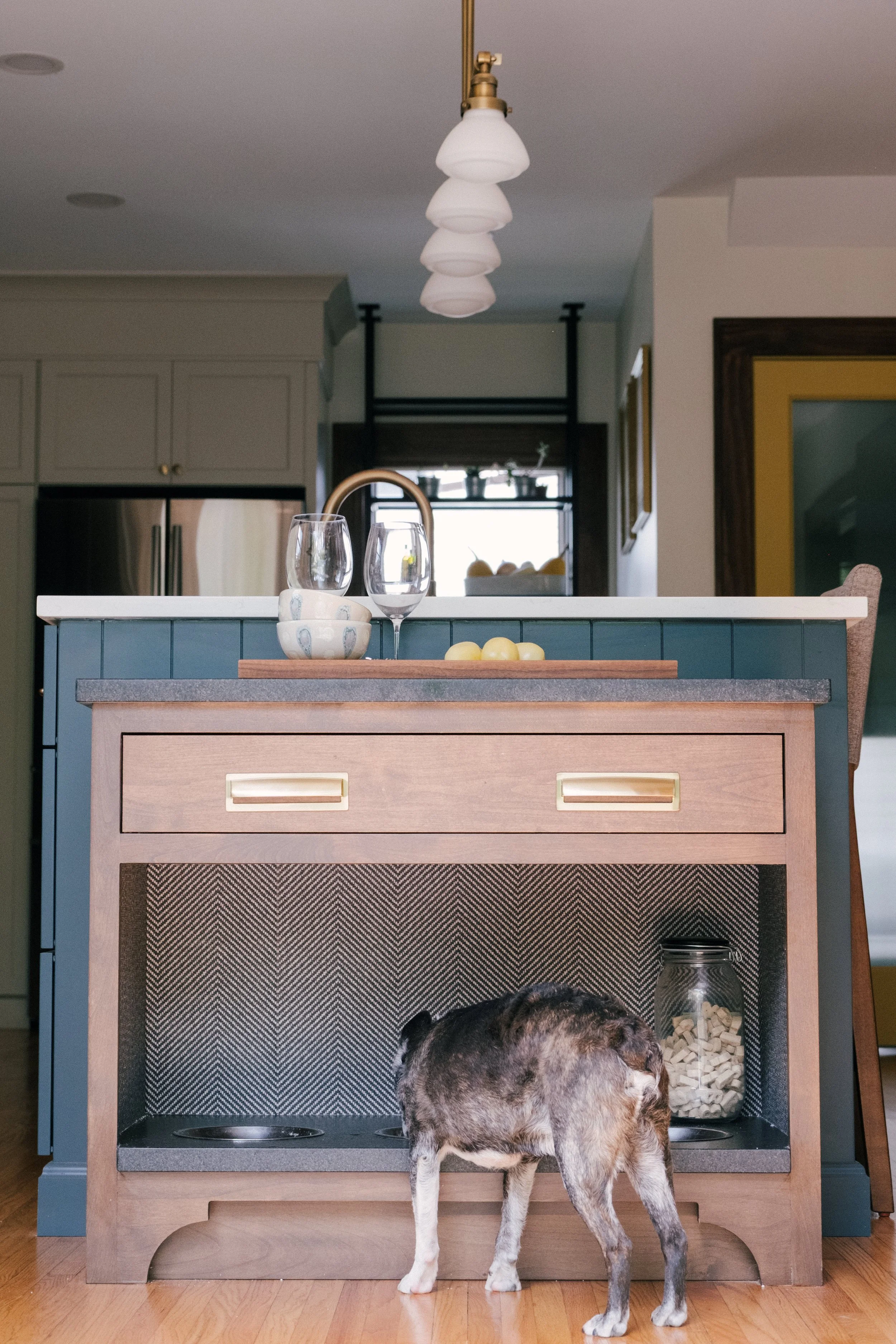 A small dog eats from a bowl in a built-in dog station just off of the kitchen island with a herringbone vinyl wallpapered backsplash. Above, a countertop holds wine glasses. small bowls and lemons.